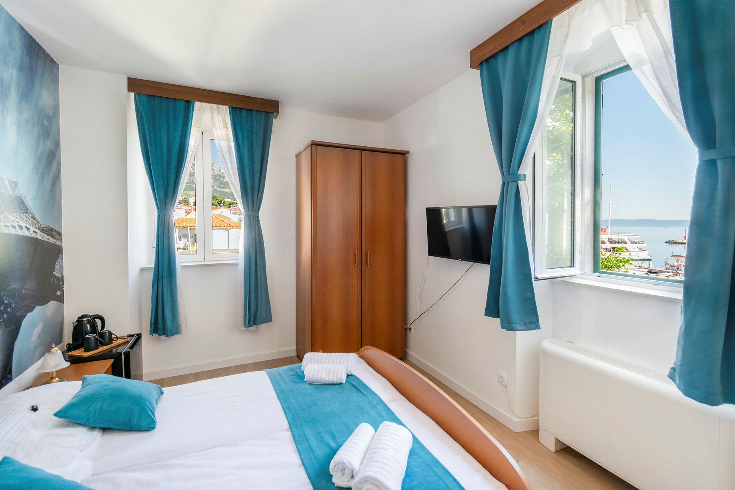 Hotelværelse med dobbeltseng, blå gardiner og udsigt til en havn udenfor vinduet. (AI-genereret tekst)