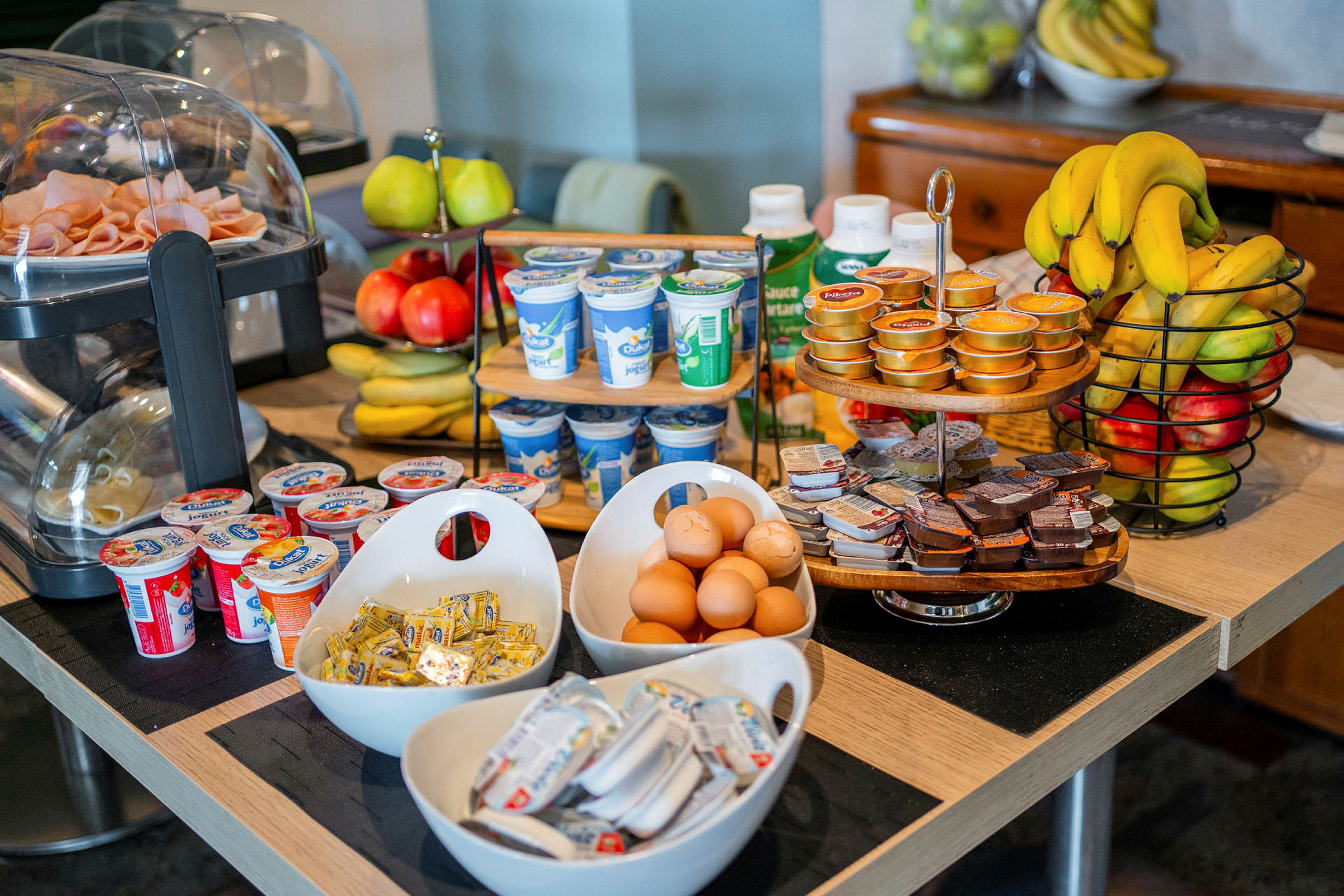 Billedet viser et morgenmadsbord med forskellige madvarer såsom æg, yoghurt, frugt, pålæg og smørpakker. (AI-genereret tekst)