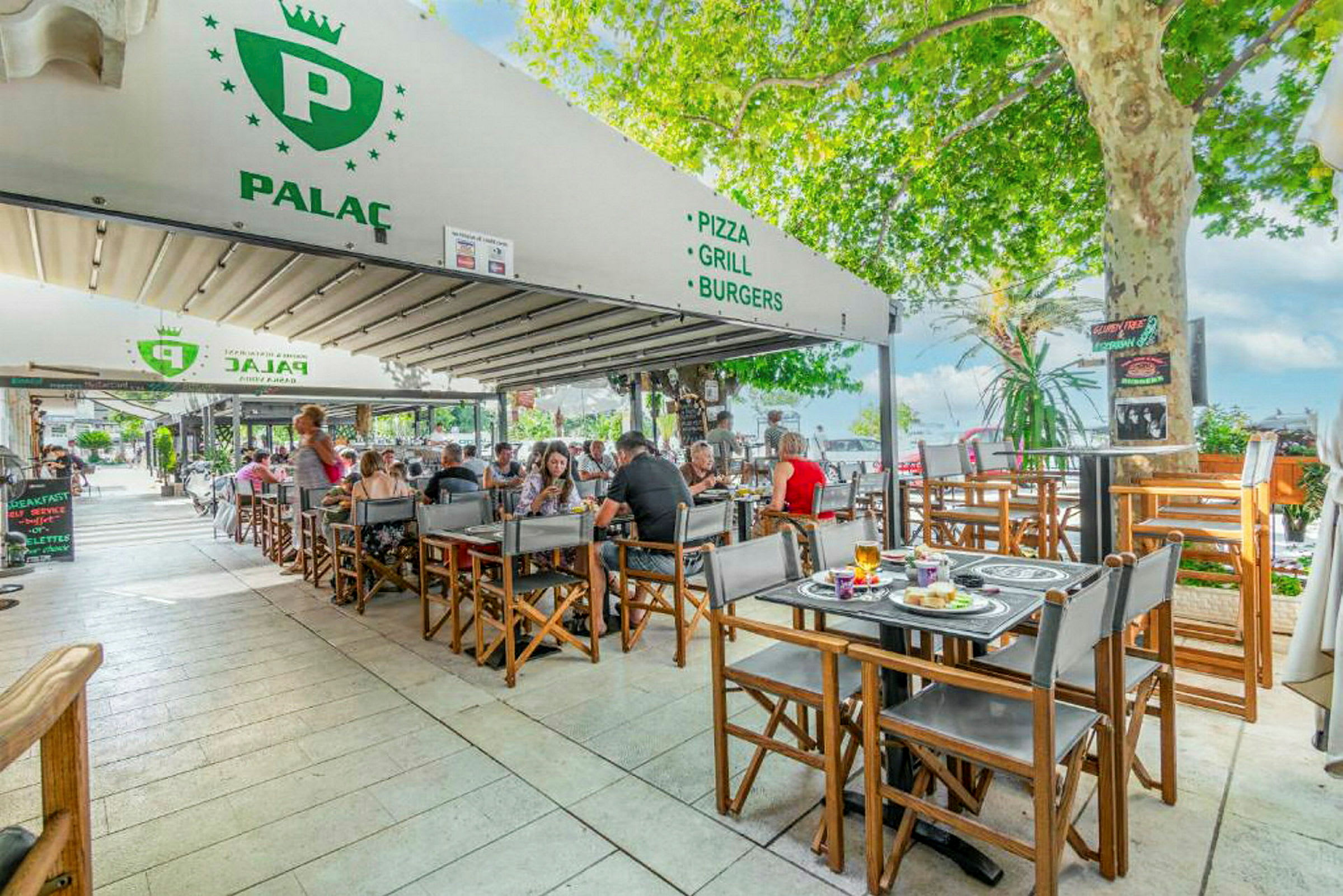 Udendørs serveringsområde ved restaurant med gæster ved borde, omgivet af grønne omgivelser. (AI-genereret tekst)