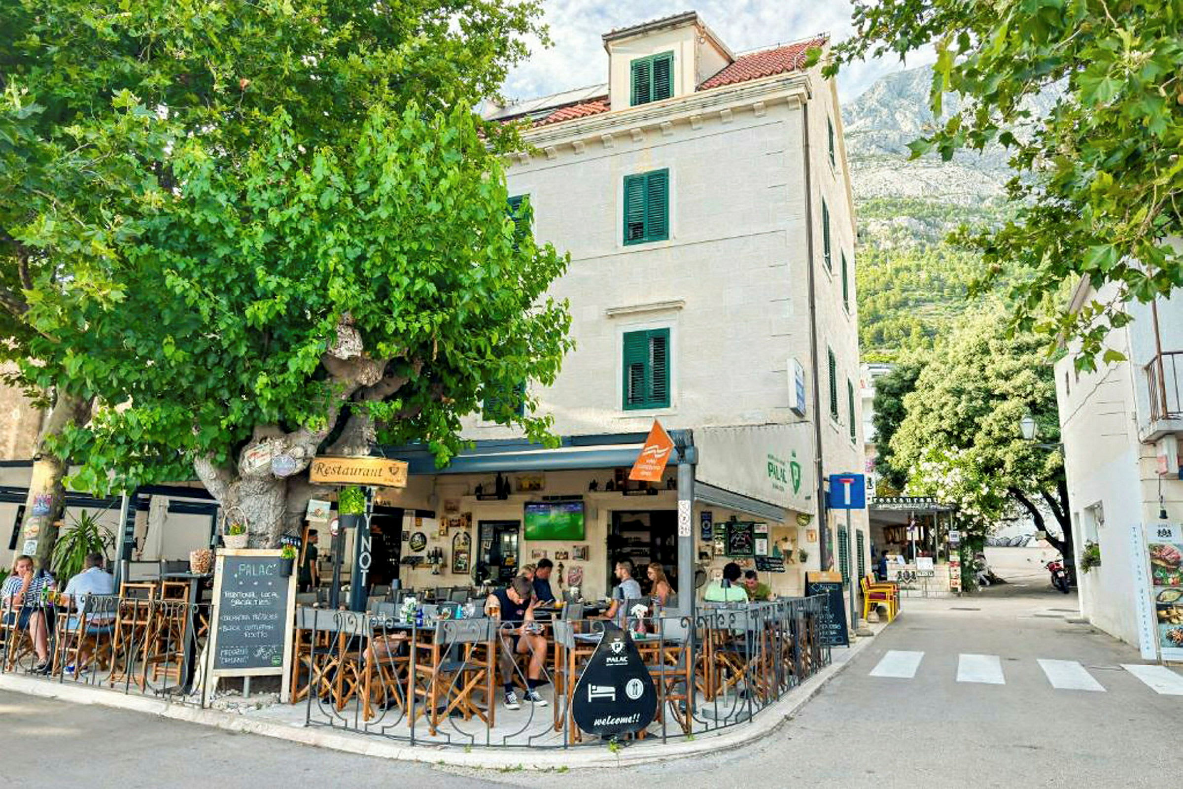 En udendørs café ved en bygning med hvide vægge og grønne skodder, omgivet af grønne områder og træer. (AI-genereret tekst)