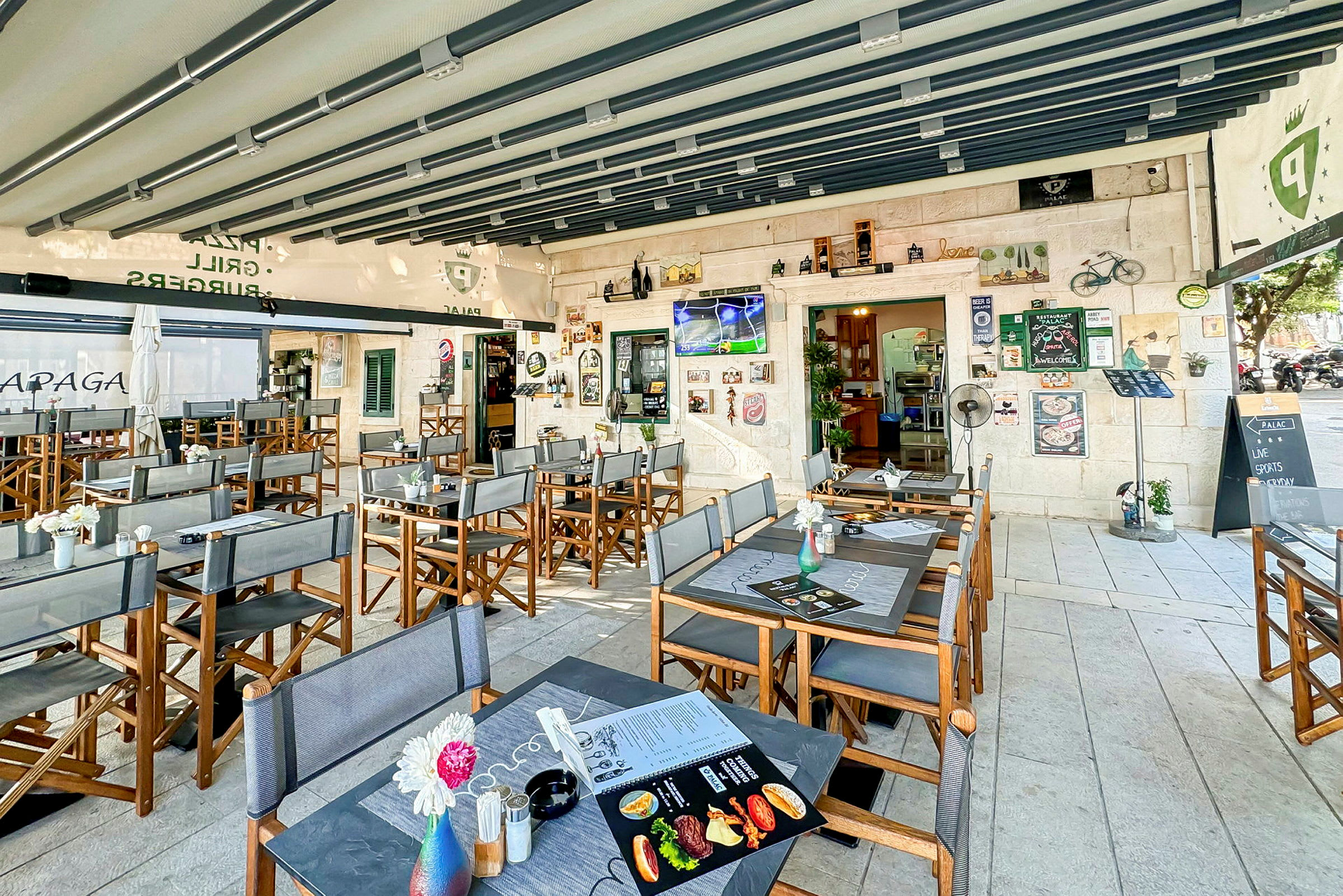 Restaurant med udendørsservering, borde og stole samt dekorationer og et TV på væggen. (AI-genereret tekst)