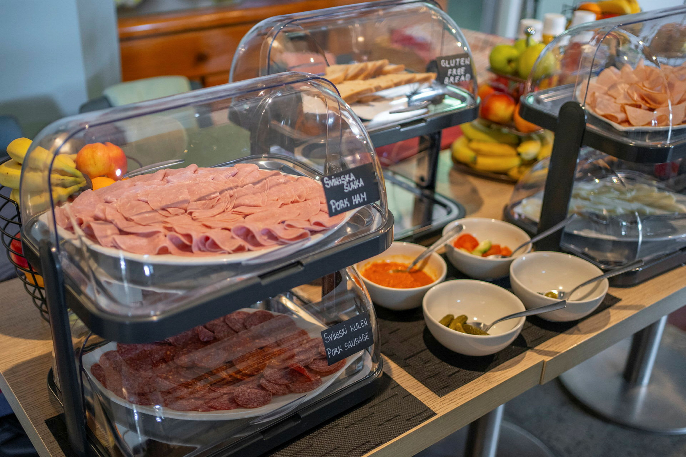 Buffetbord med skiver af skinke og pølse, et udvalg af dressinger og grøntsager samt frugt i baggrunden. (AI-genereret tekst)