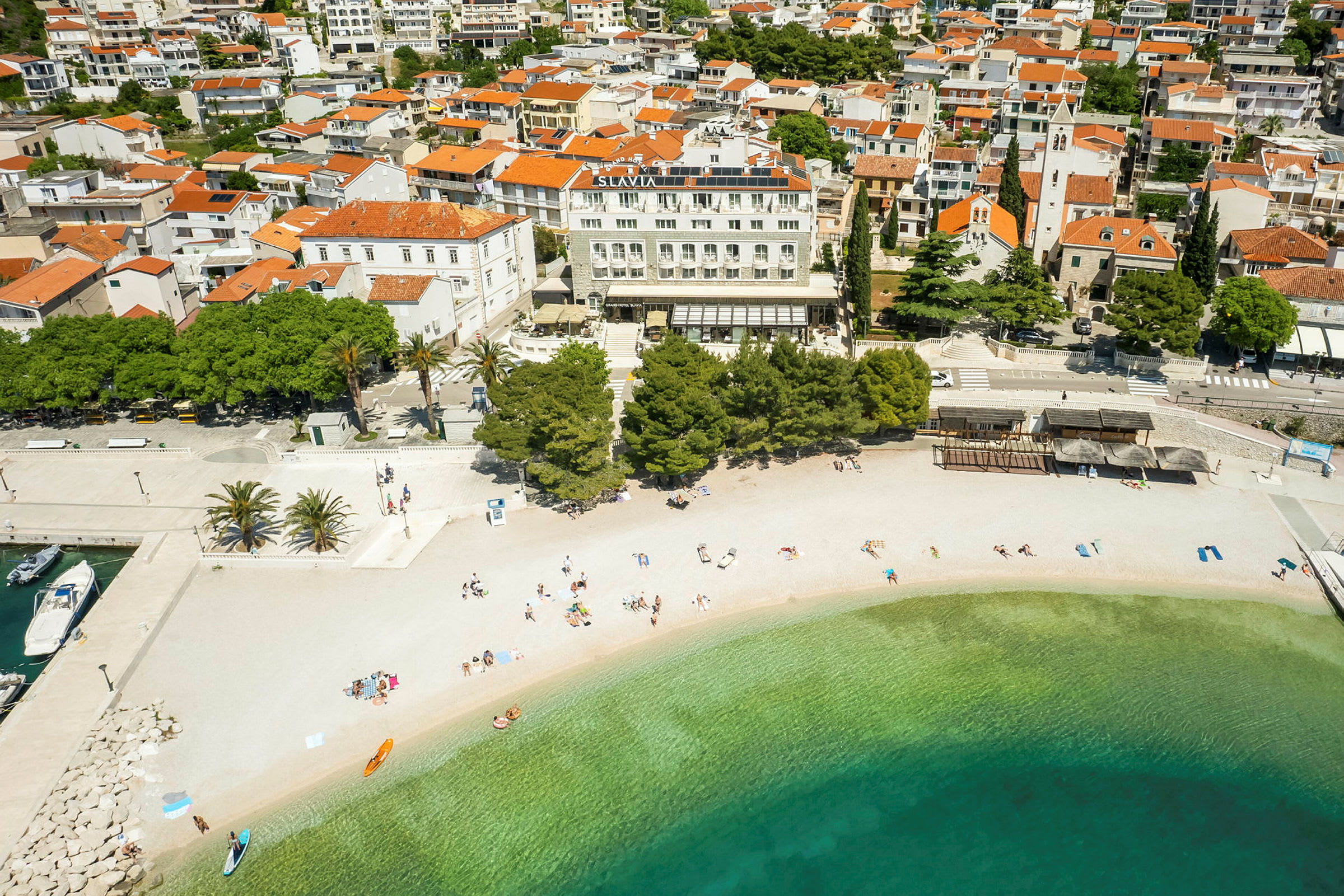 Luftfoto af en strand med klart vand foran en by med røde tegltage på huse og bygninger. (AI-genereret tekst)