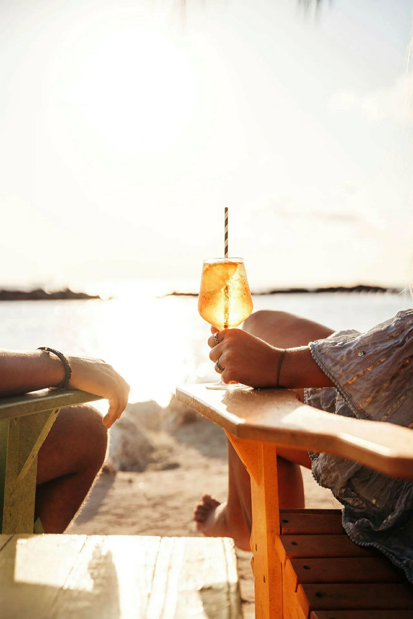To personer sidder i stole på en strand med udsigt over havet og solnedgangen. I forgrunden holdes et glas med en drink. (AI-genereret tekst)
