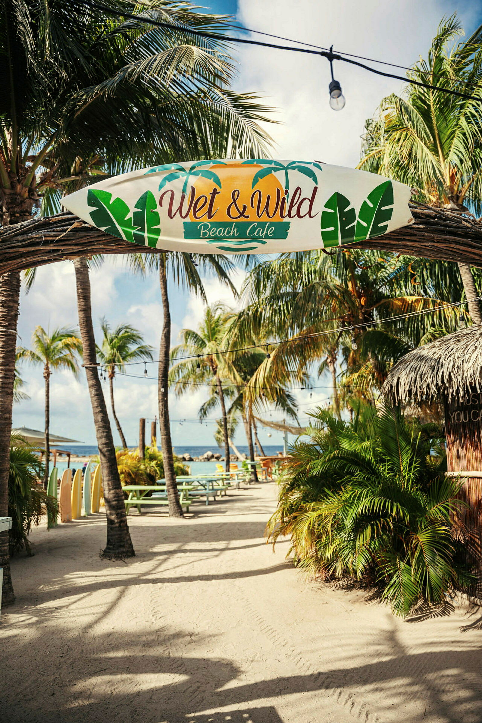 Skiltet med teksten "Wet & Wild Beach Café" hænger på en bue i et udendørs område med palmer, borde og hav i baggrunden. (AI-genereret tekst)