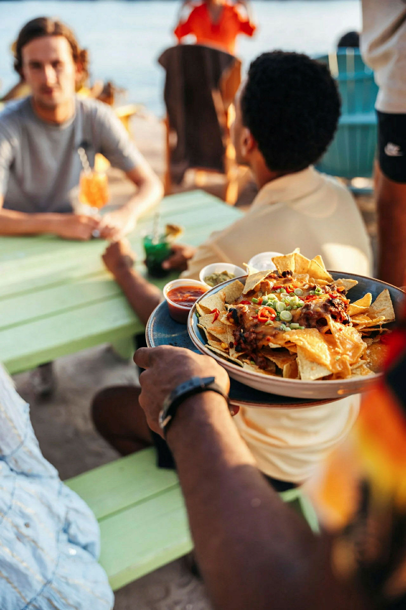 En person holder en tallerken med nachos, hvor forskellige dipsaucer er placeret rundt om tallerkenen. Scenen er udendørs ved et bord nær vand. (AI-genereret tekst)