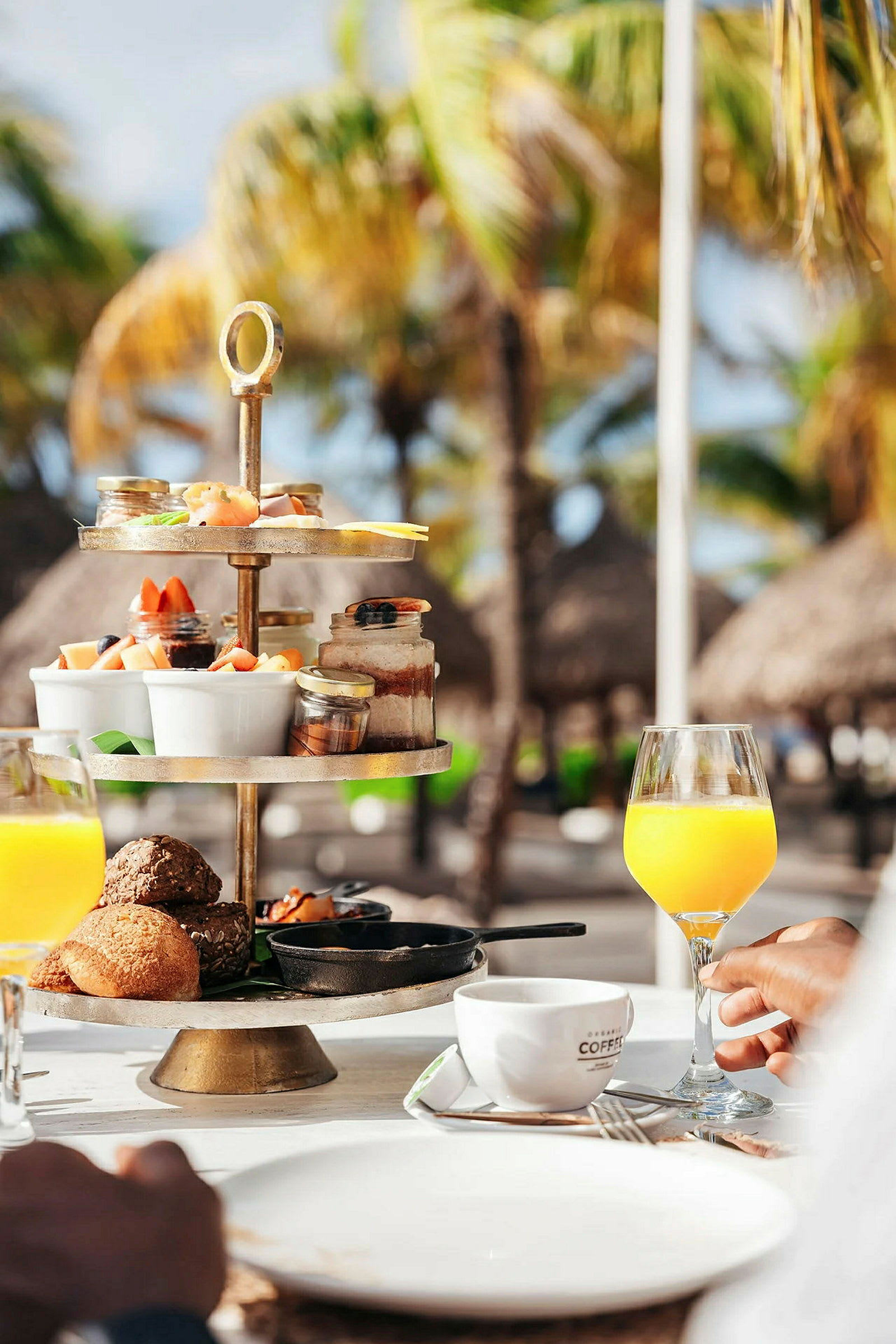 Et morgenbord dækket med et kagefad med forskellige små portioner mad, et glas med appelsinjuice og en kaffekop i forgrunden. I baggrunden ses palmer og tropiske parasoller. (AI-genereret tekst)