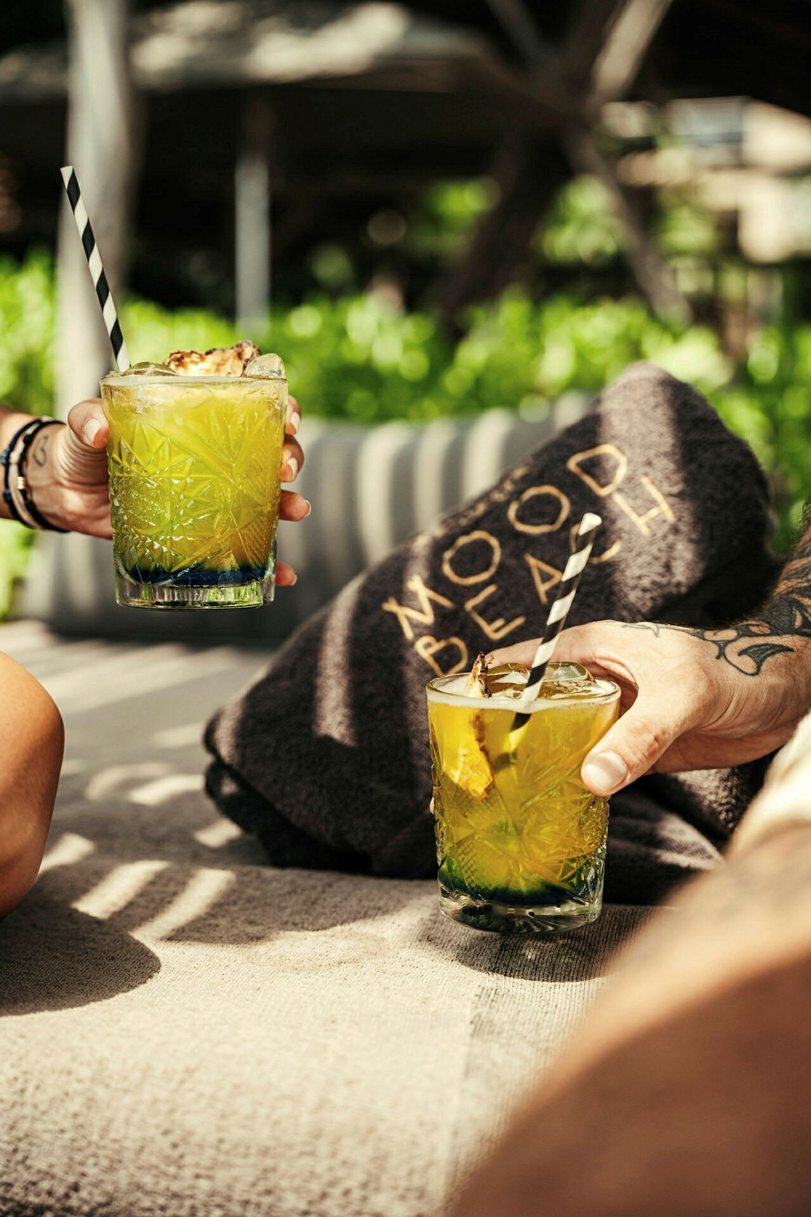 To hænder holder glas med grønne drinks og sort-hvide sugerør på solsenge med et tæppe mærket 'Mood Beach' i baggrunden. (AI-genereret tekst)