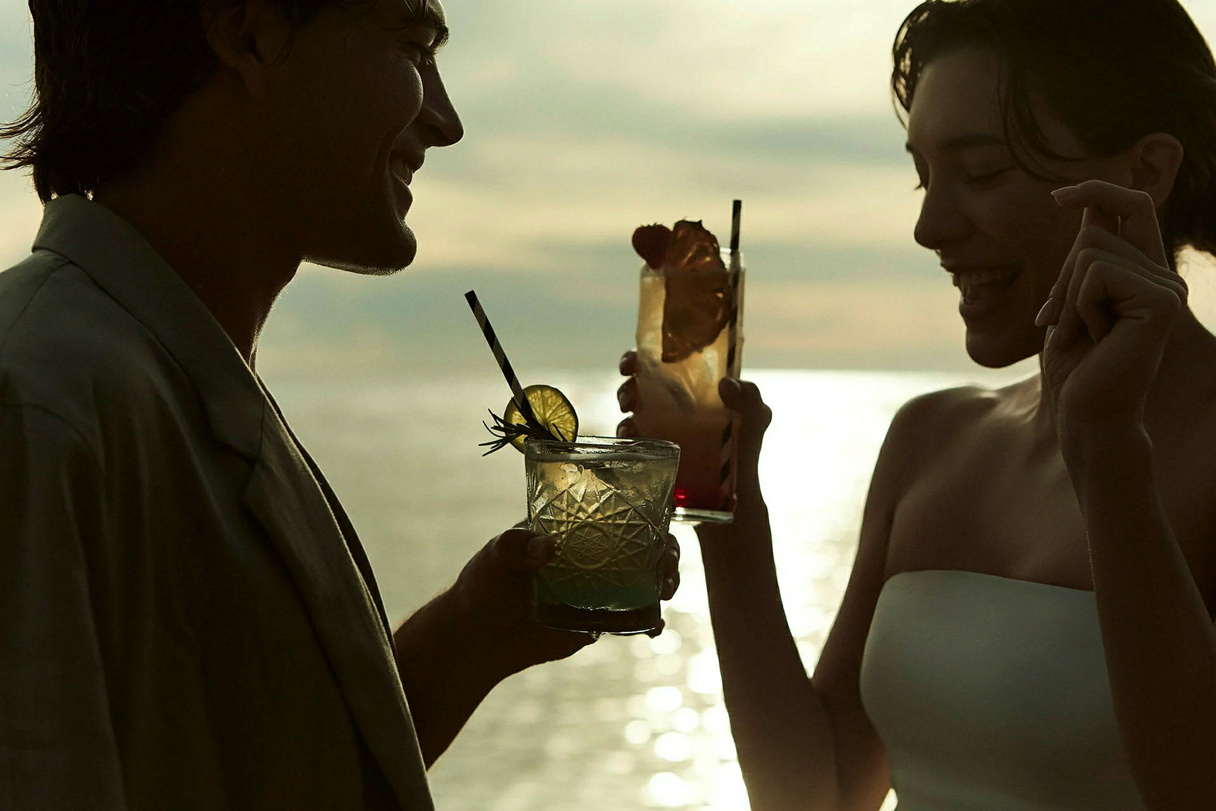 To personer holder glas med drinks mod en solnedgang. (AI-genereret tekst)