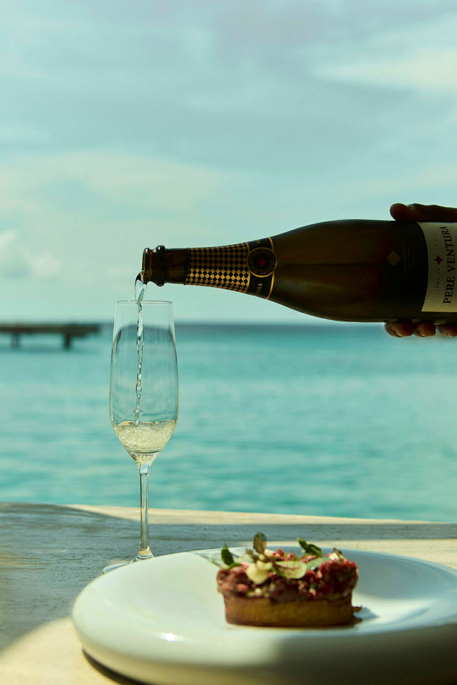Et glas fyldes med champagne ved siden af en tallerken med en lille dessert, med havudsigt i baggrunden. (AI-genereret tekst)