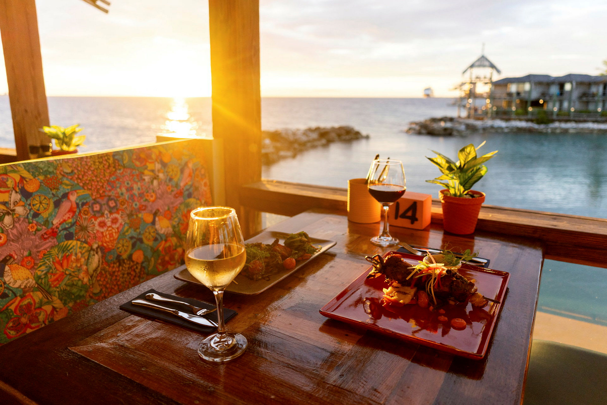 En restaurantstolsryg med farverigt mønster, et velanrettet bord med vinglas og mad, udsigt over havet i solnedgangen. (AI-genereret tekst)