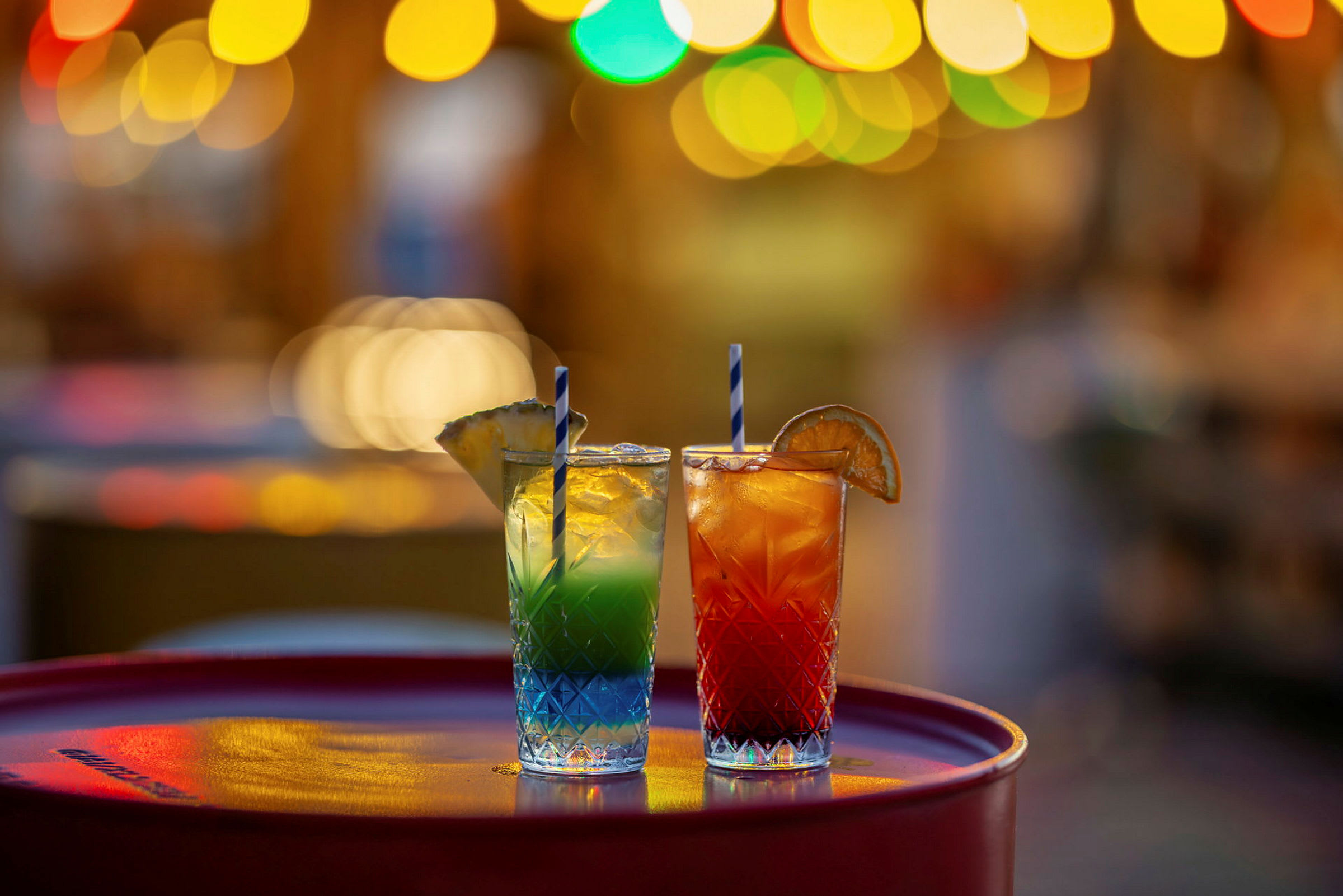 To glas med farverige cocktails, pyntet med frugt, står på et bord med uskarpe lys i baggrunden. (AI-genereret tekst)