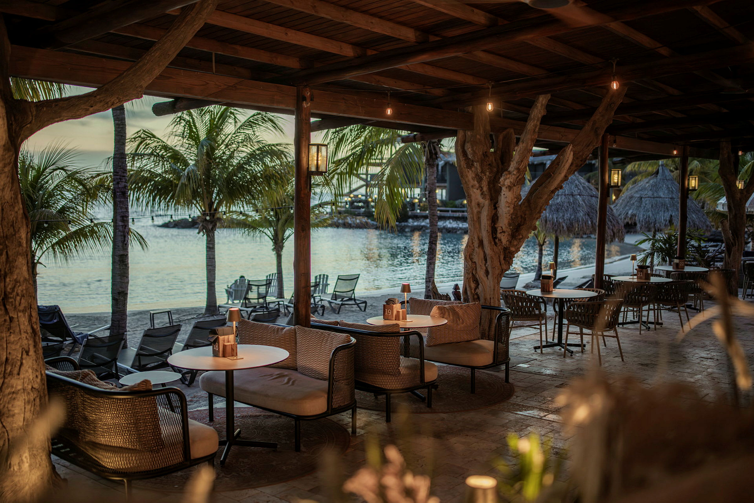En udendørs lounge under et skur af træ med sofaer og borde ved havet, omgivet af palmetræer. (AI-genereret tekst)
