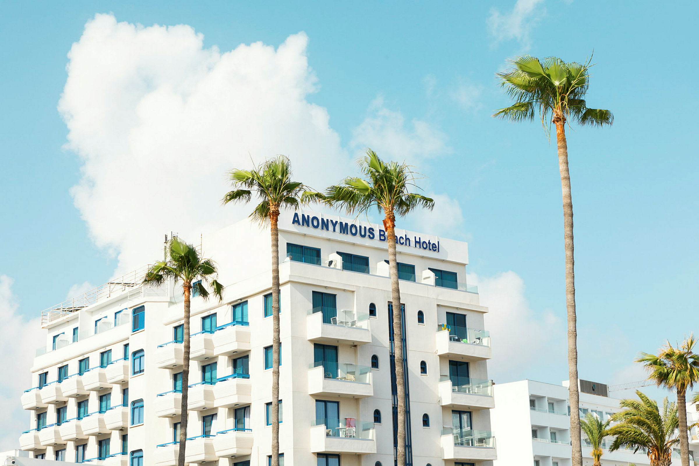 Et hotel med navnet Anonymous Beach Hotel, hvid facade og blå vinduer, omgivet af palmer. (AI-genereret tekst)