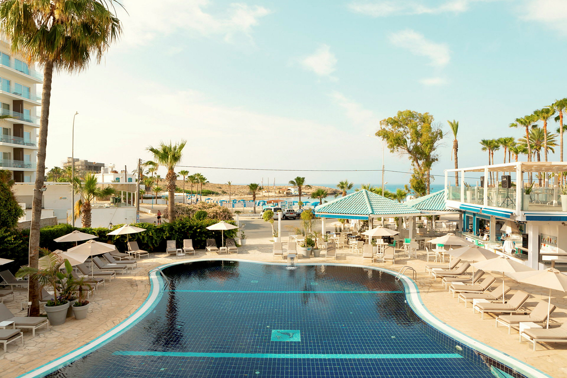Hotelområde med pool, liggestole, parasoller og palmer med strand og hav i baggrunden. (AI-genereret tekst)