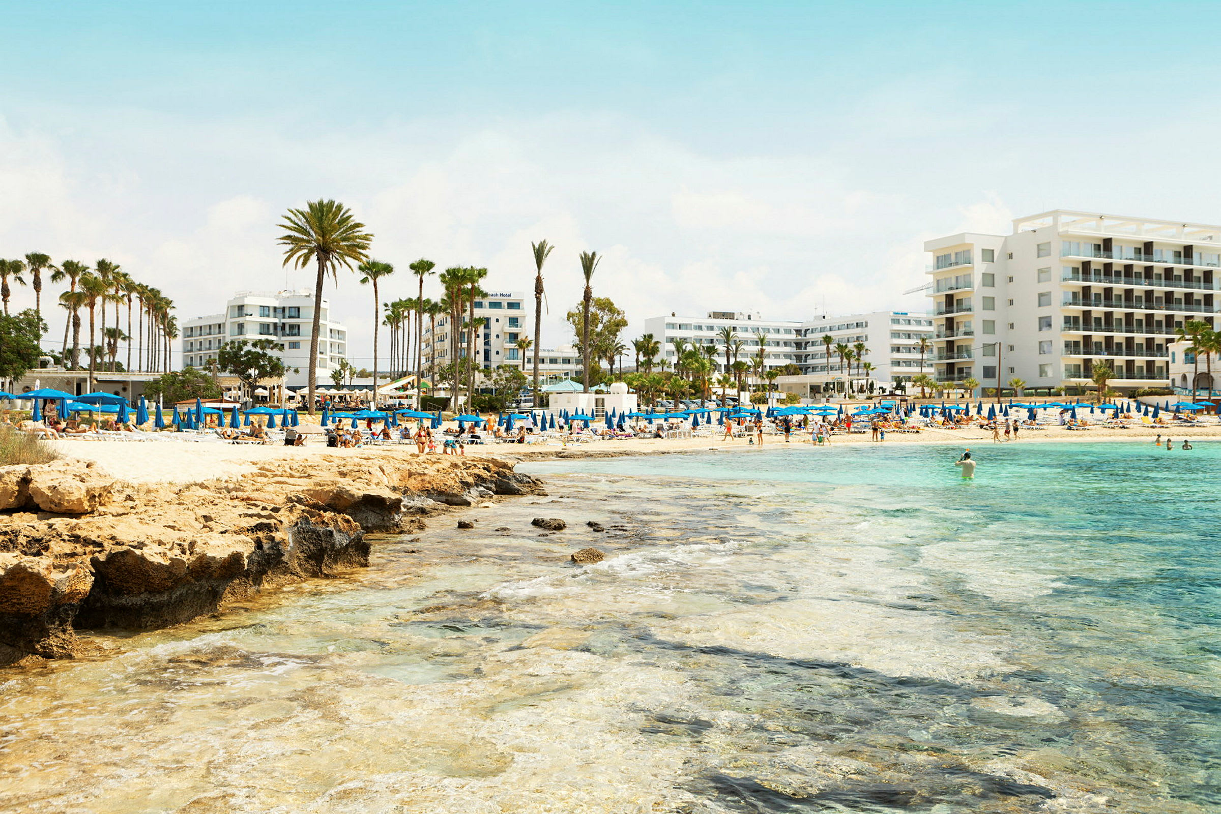 Strand med blå parasoller, palmer og hotelbygninger i baggrunden. (AI-genereret tekst)