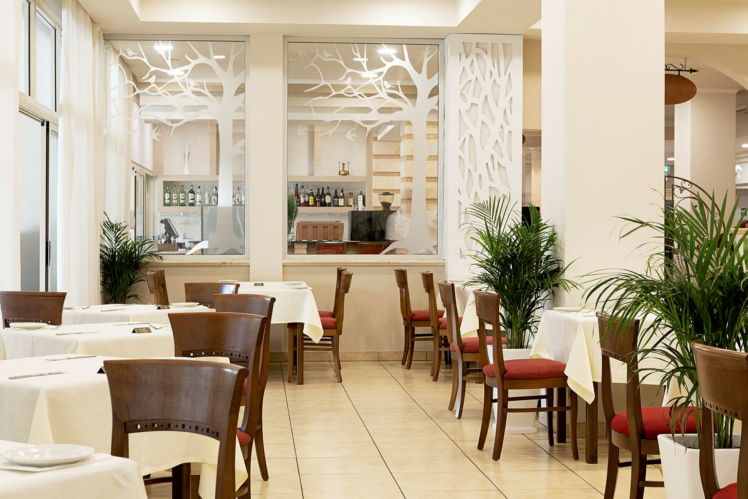 En restaurant med borde og stole, dekoreret med planter og en glasvæg med et træsdesignmotiv. (AI-genereret tekst)