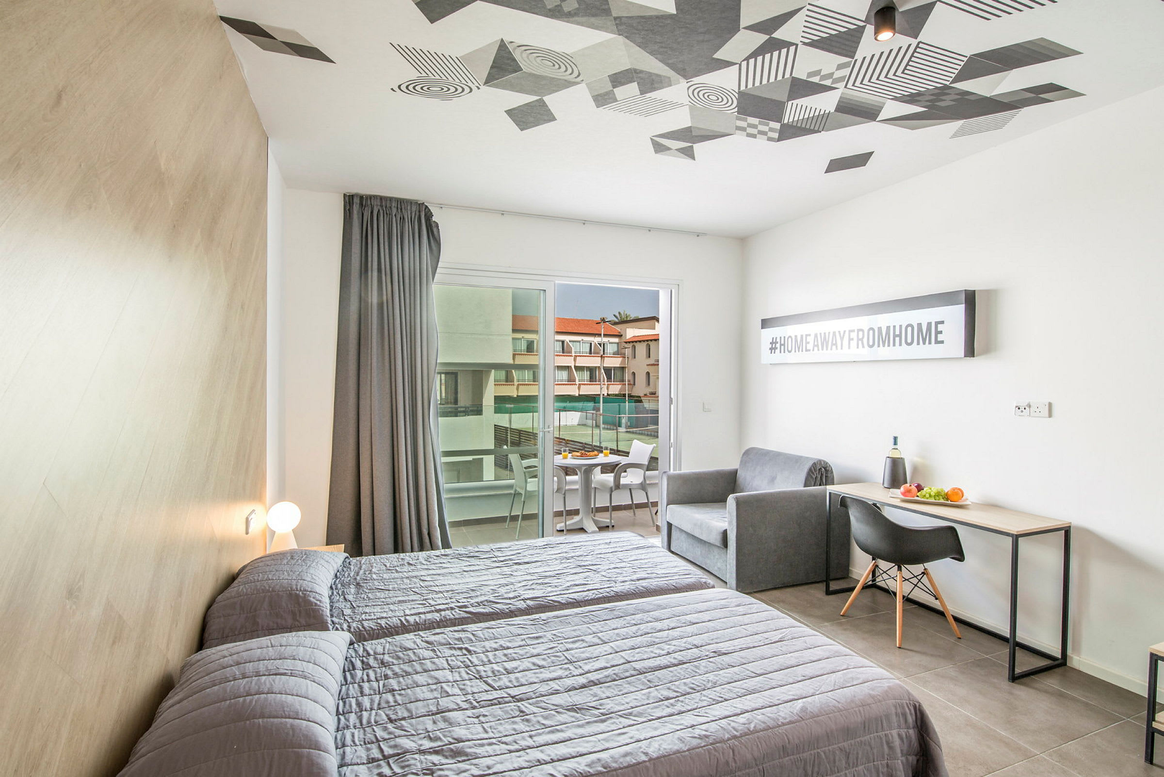 Et hotelværelse med to senge, en sofa, et skrivebord og en altan med udsigt til en bygning. (AI-genereret tekst)