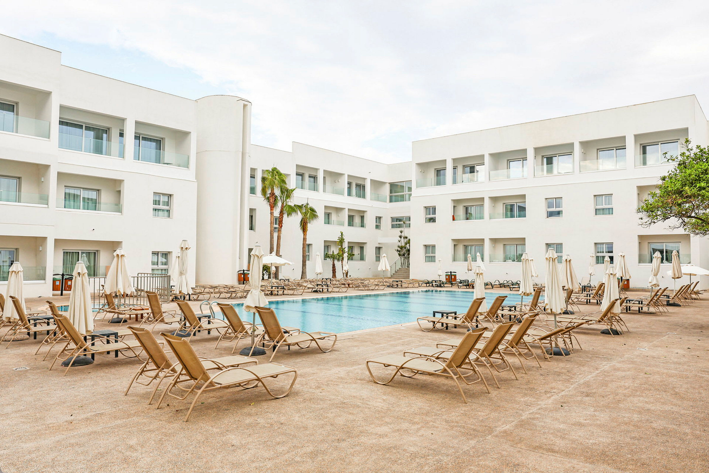 Udendørs pool omgivet af liggestole og hvide bygninger i et hotelområde. (AI-genereret tekst)