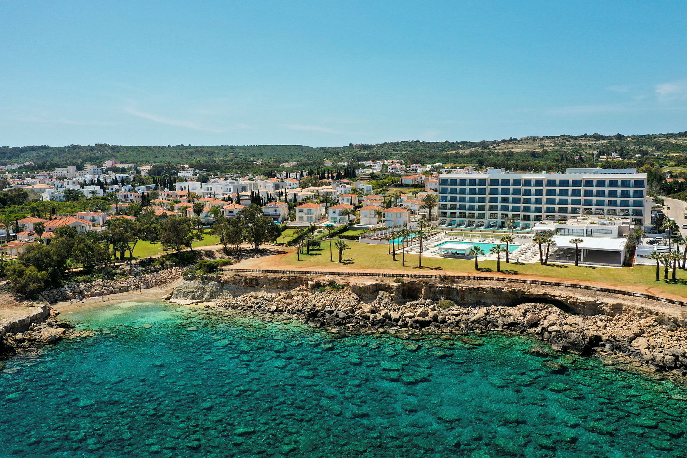 En kystlinje med klart turkis vand, en klippe og bygninger omkring et hotel med poolområde. (AI-genereret tekst)