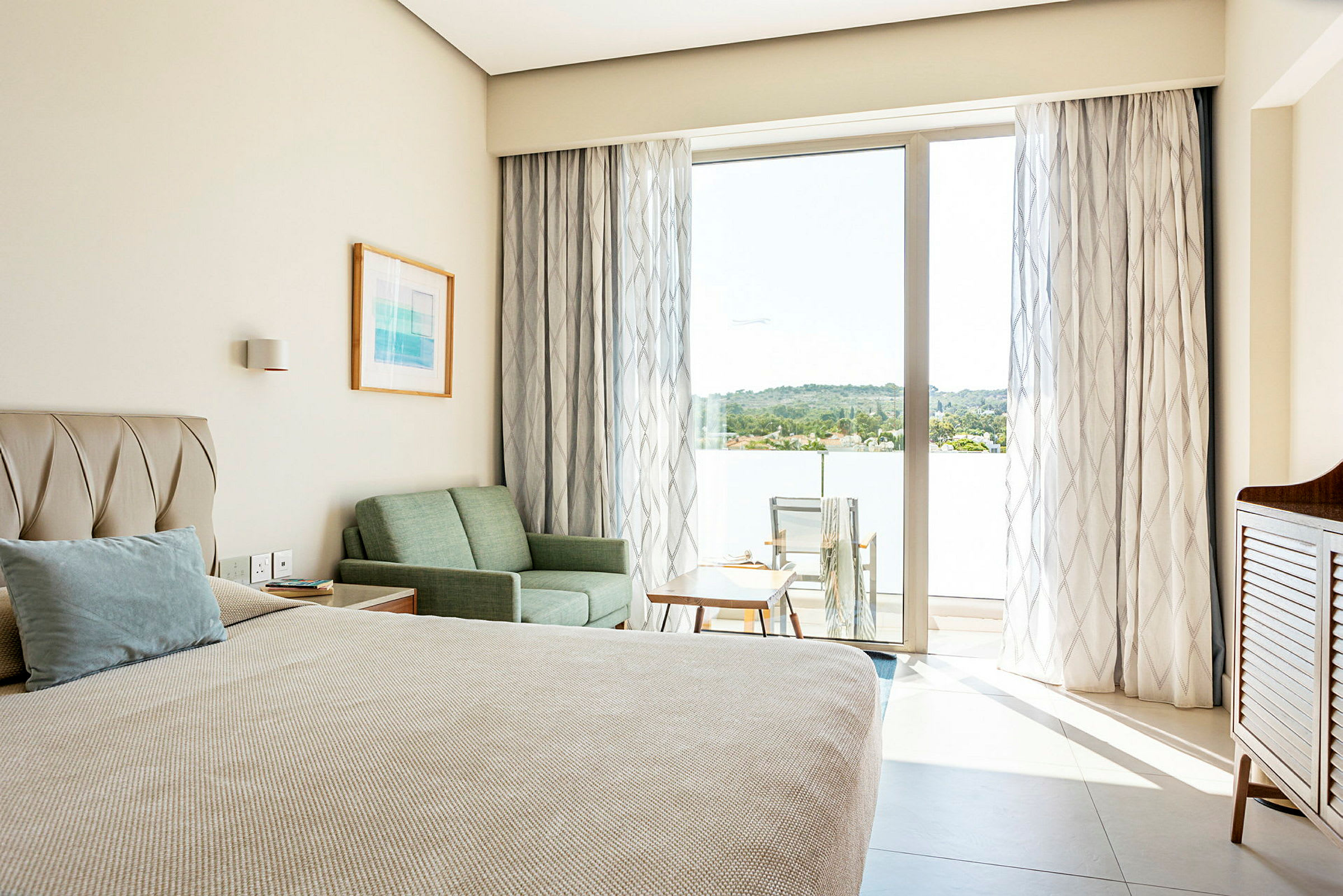 Et hotelværelse med en seng, sofa og bord samt en balkon med stole og udsigt over grønne områder. (AI-genereret tekst)