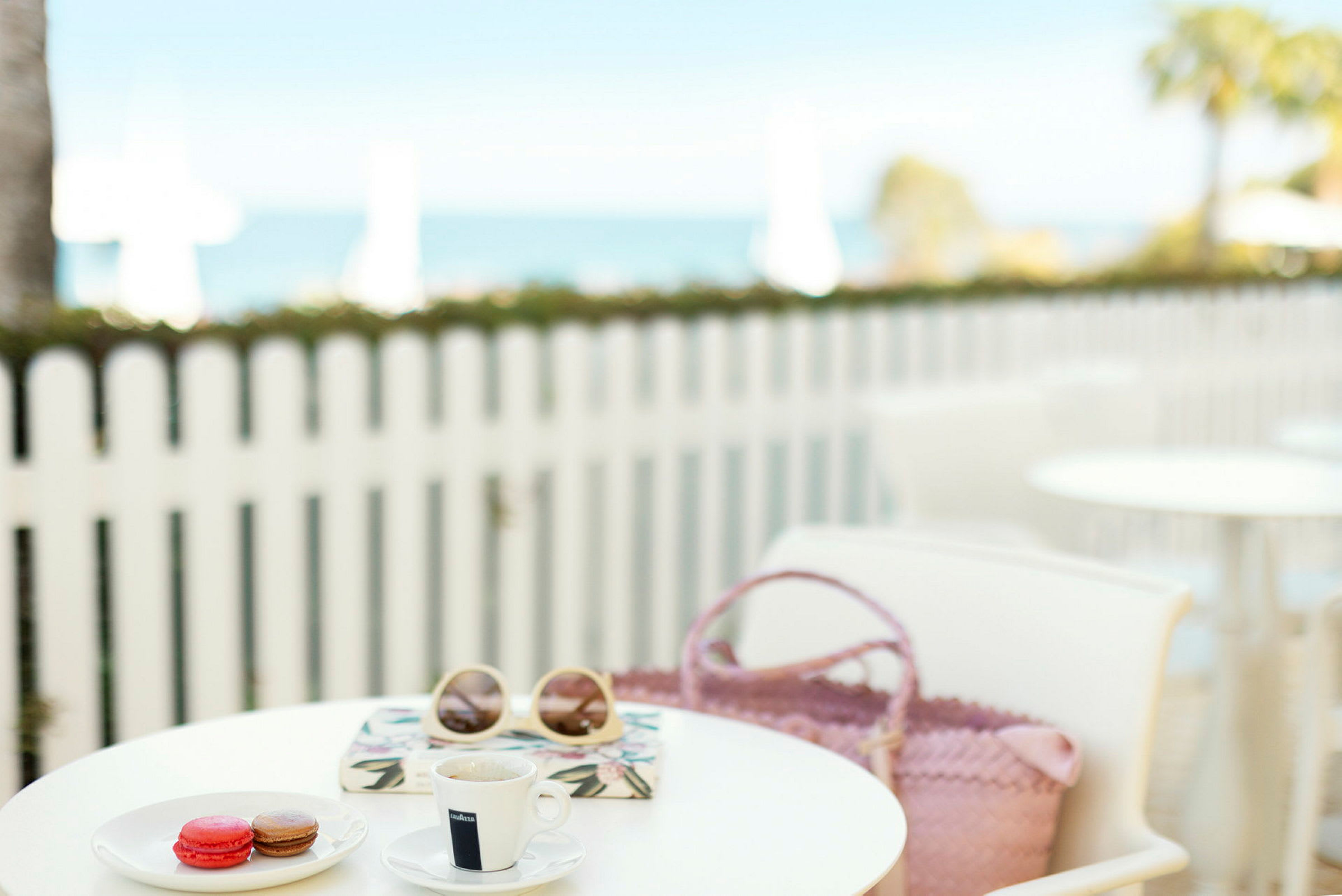 En kop kaffe, macarons, solbriller og en taske på et cafébord med et hvidt træhegn og havet i baggrunden. (AI-genereret tekst)