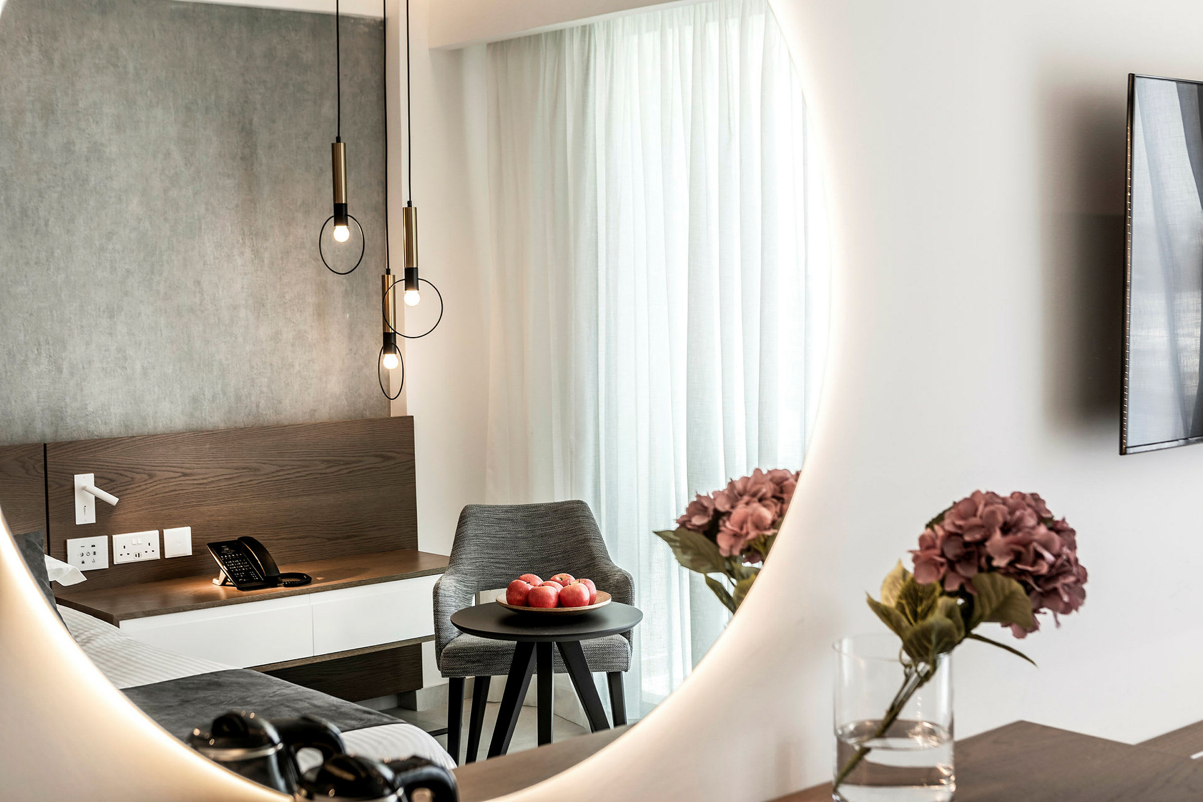 Interiør af hotelværelse med et spejl, der reflekterer en seng, et natbord med en telefon, en skål med røde æbler på et rundt bord og en vase med lilla blomster. (AI-genereret tekst)