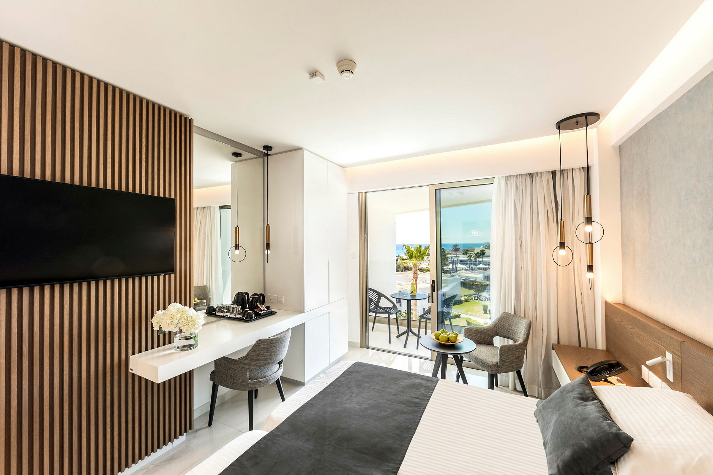Moderne hotelværelse med en dobbeltseng, bord og stole samt udsigt over havet fra en balkon. (AI-genereret tekst)