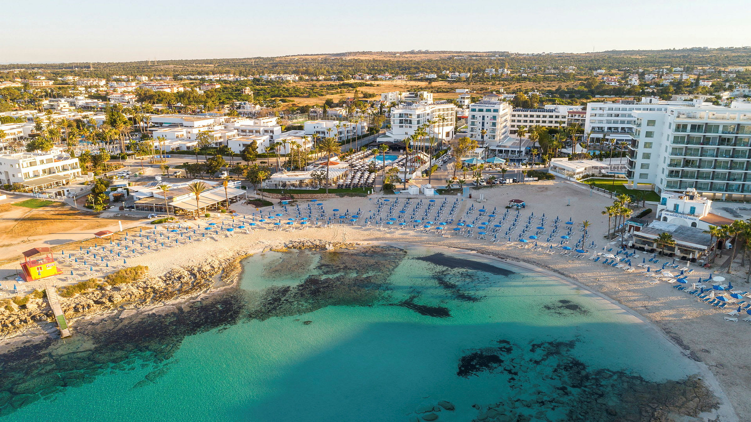 Strand med parasoller og stole, omgivet af turkis vand, bygninger og grønne områder. (AI-genereret tekst)