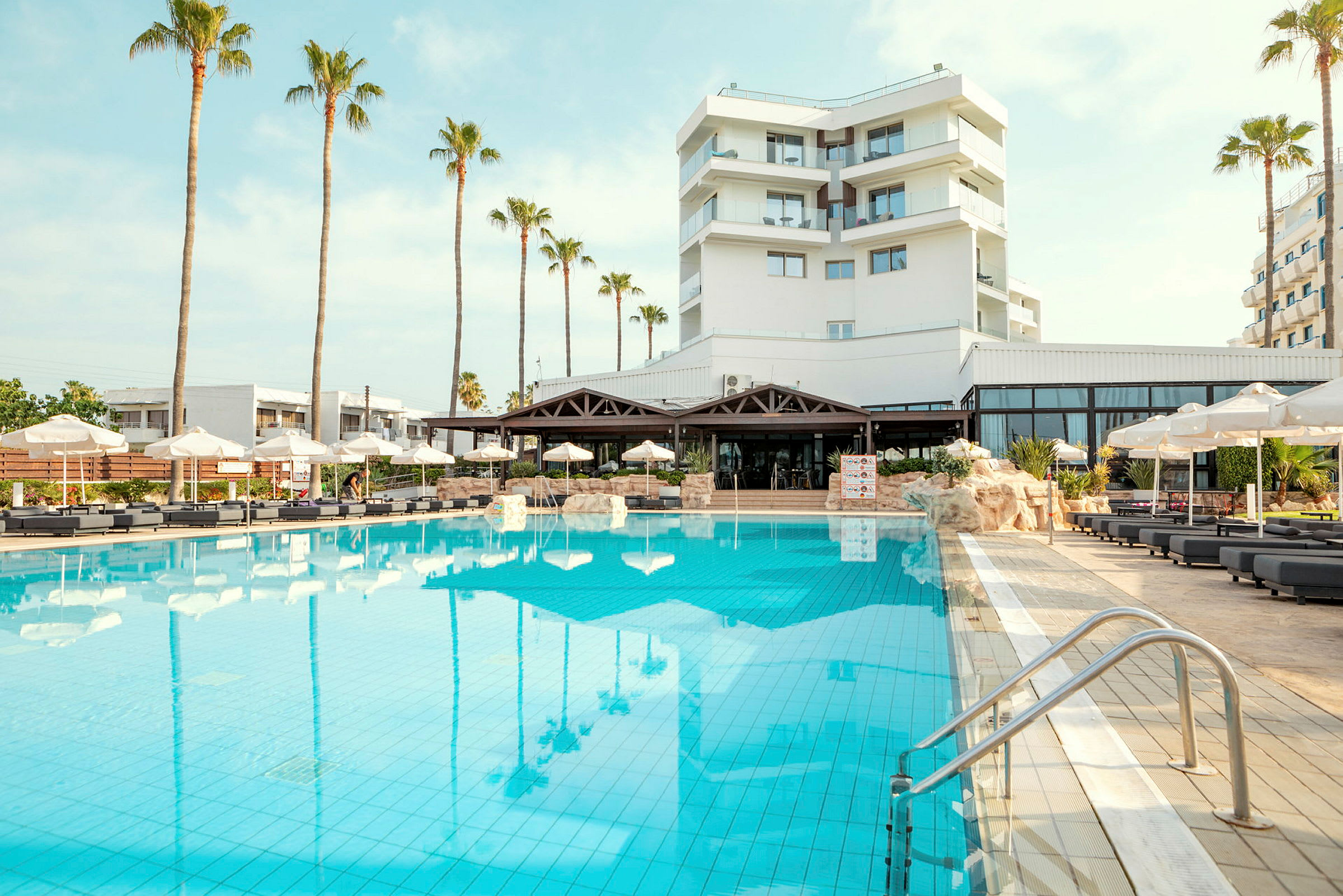 Billede af hotelbygning med en stor udendørs pool og liggestole under parasoller. (AI-genereret tekst)