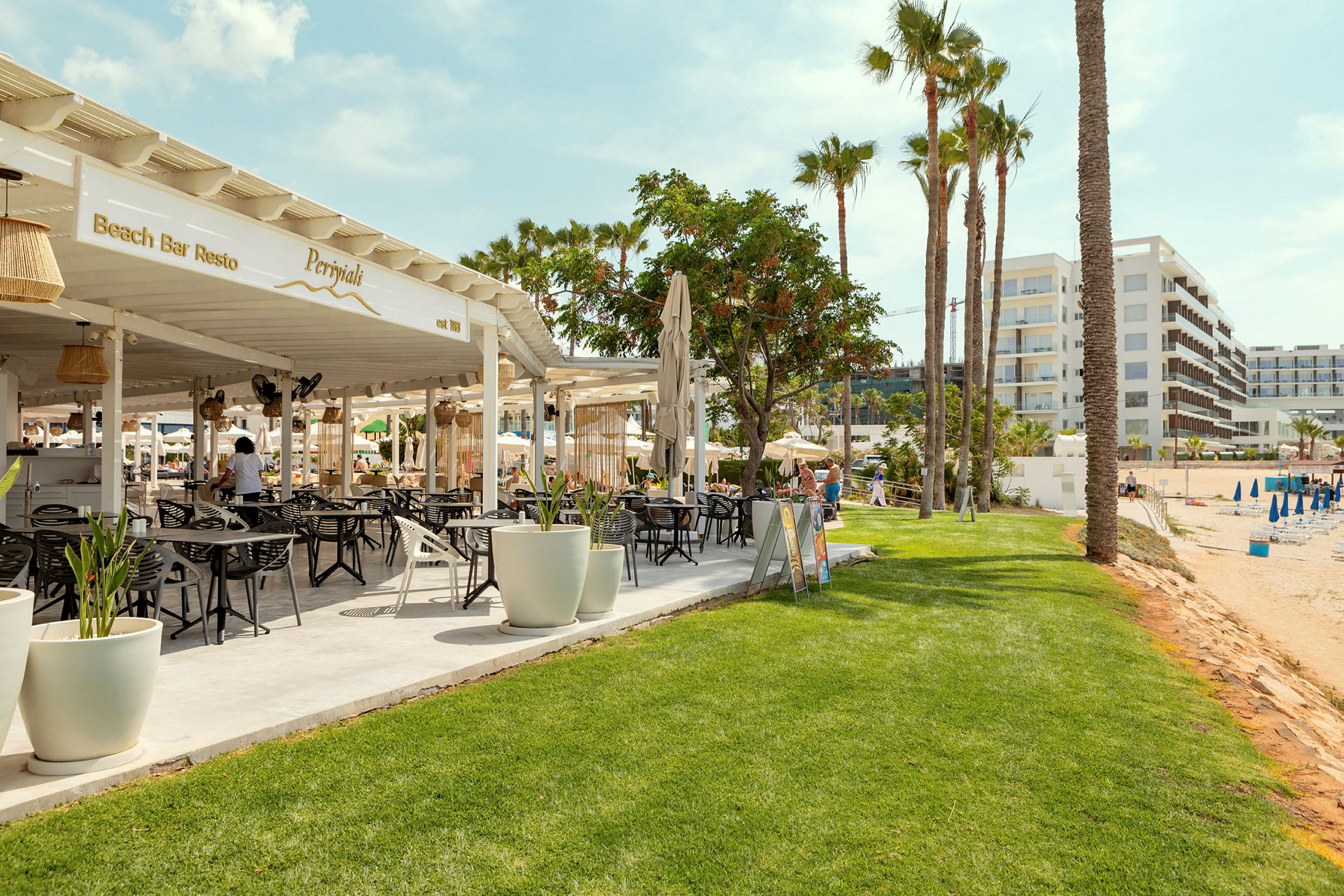 Billede af en strandbar med siddepladser og parasoller samt hotelbygninger i baggrunden. (AI-genereret tekst)