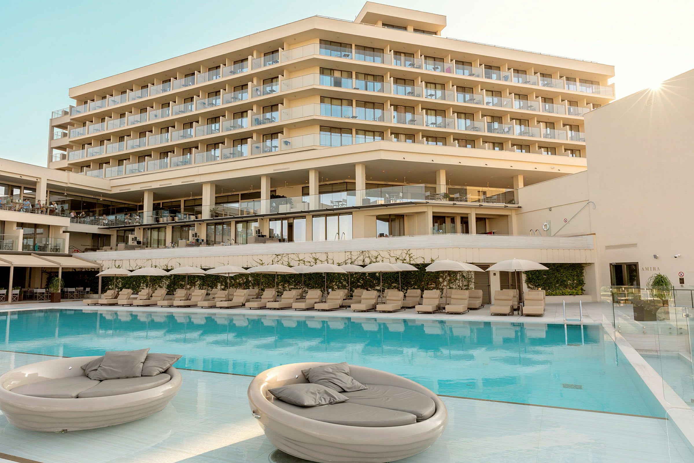 Et hotel med flere etager og store altaner ses bag en swimmingpool med solsenge og parasoller. (AI-genereret tekst)