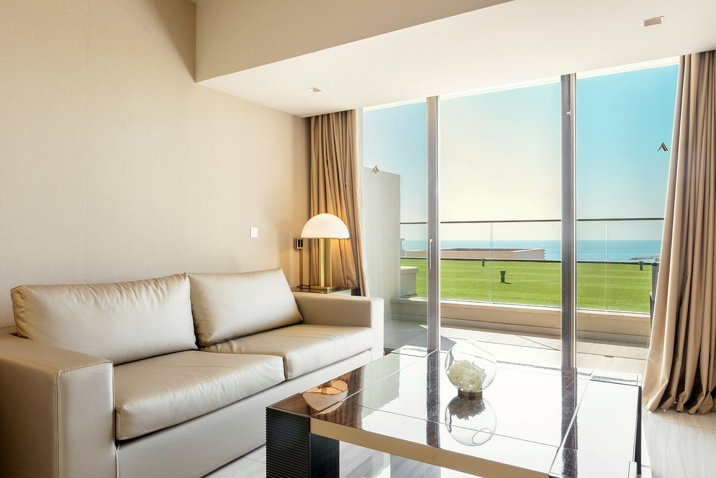Billede af en moderne stue med en beige sofa, glasbord og en stor vinduesdør, der åbner ud til en balkon med udsigt mod havet. (AI-genereret tekst)