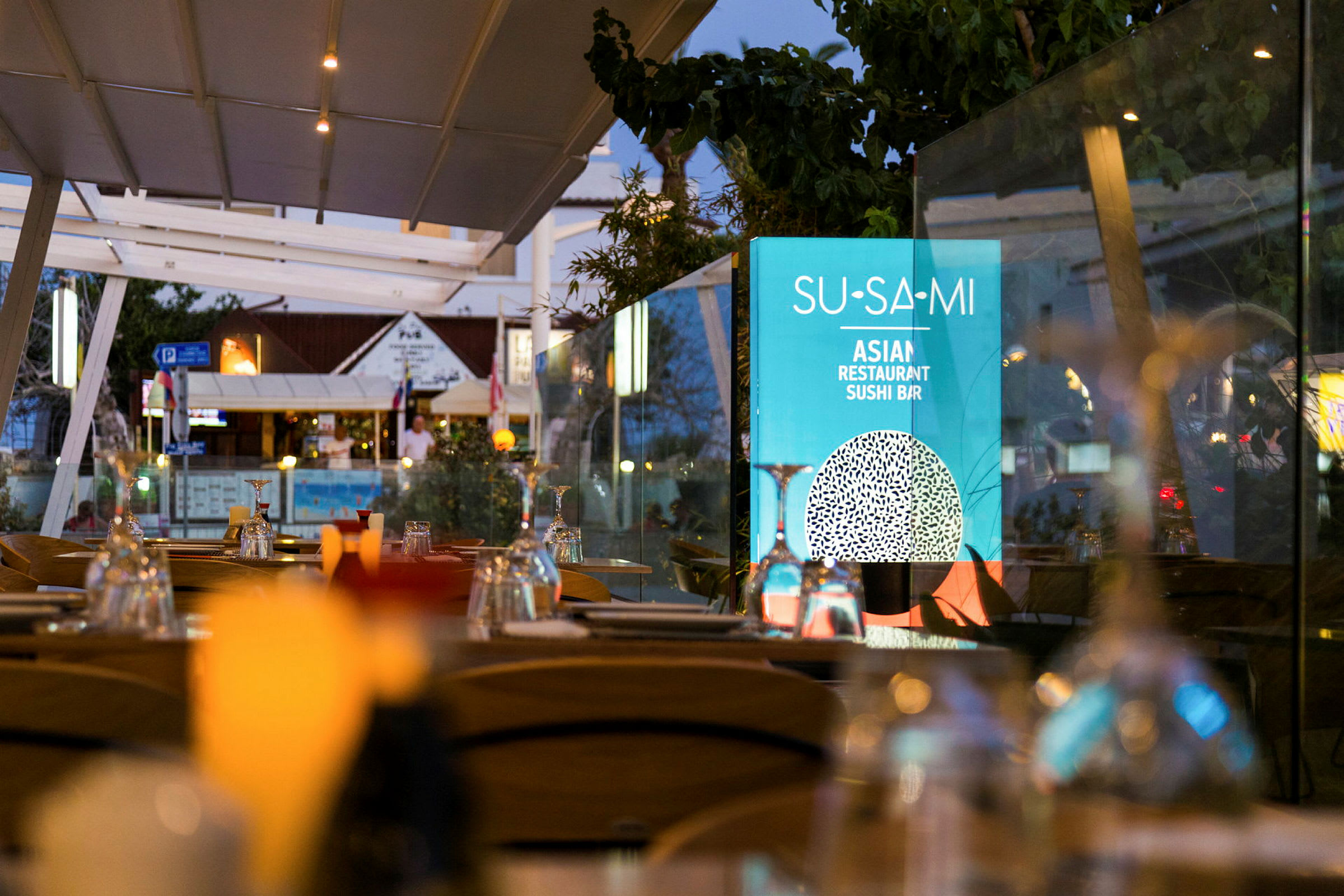 Udendørs serveringsområde med dækkede borde og et skilt med teksten 'Su-Sa-Mi Asian Restaurant Sushi Bar'. (AI-genereret tekst)