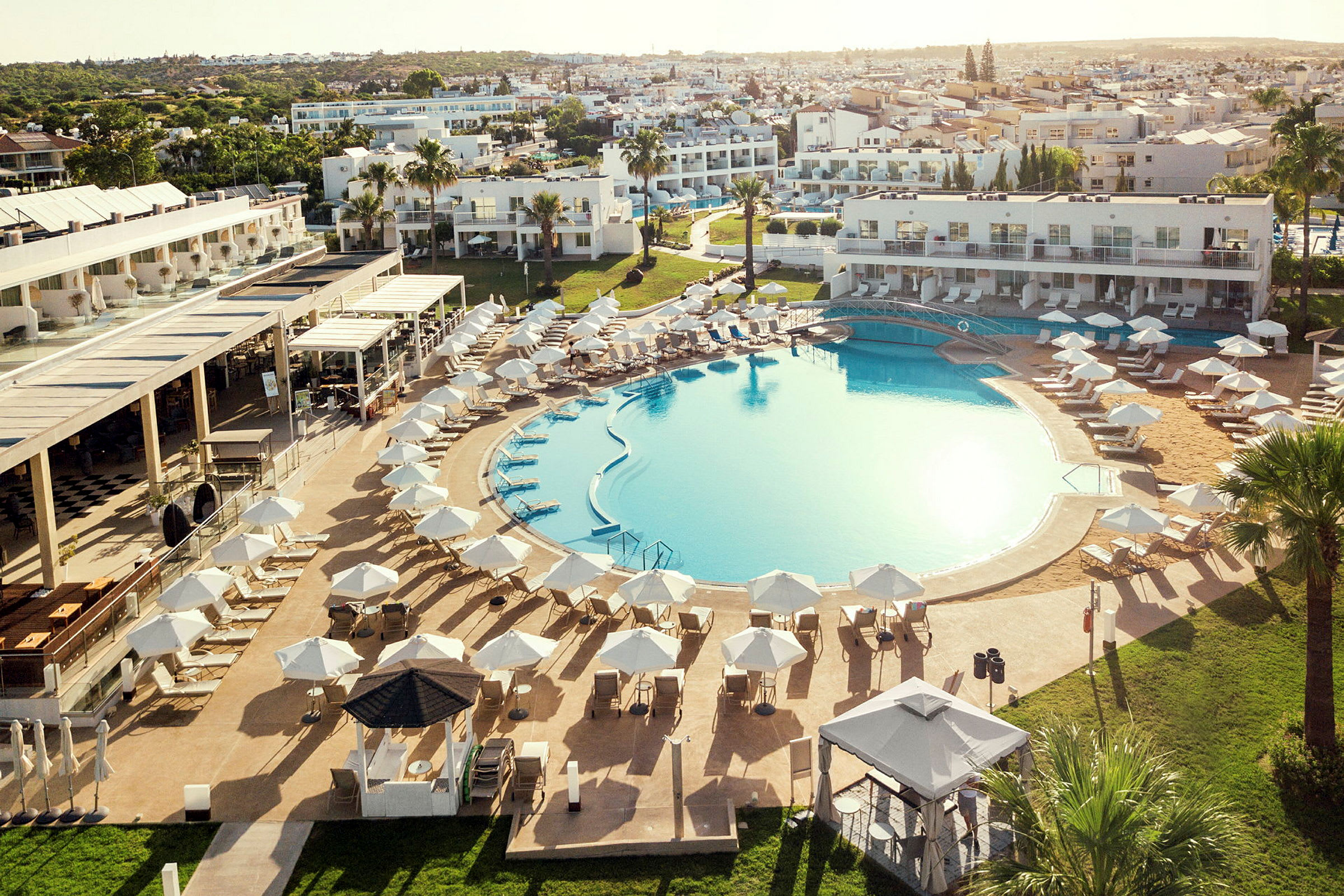 En stor udendørs pool omgivet af liggestole og parasoller, med hotelbygninger i baggrunden. (AI-genereret tekst)