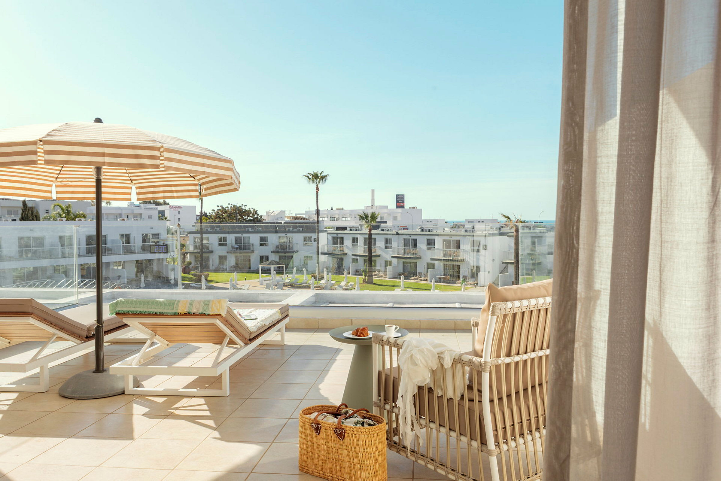 En solrig terrasse med liggestole, parasol, udsigt over en bygning og noget grønt. (AI-genereret tekst)