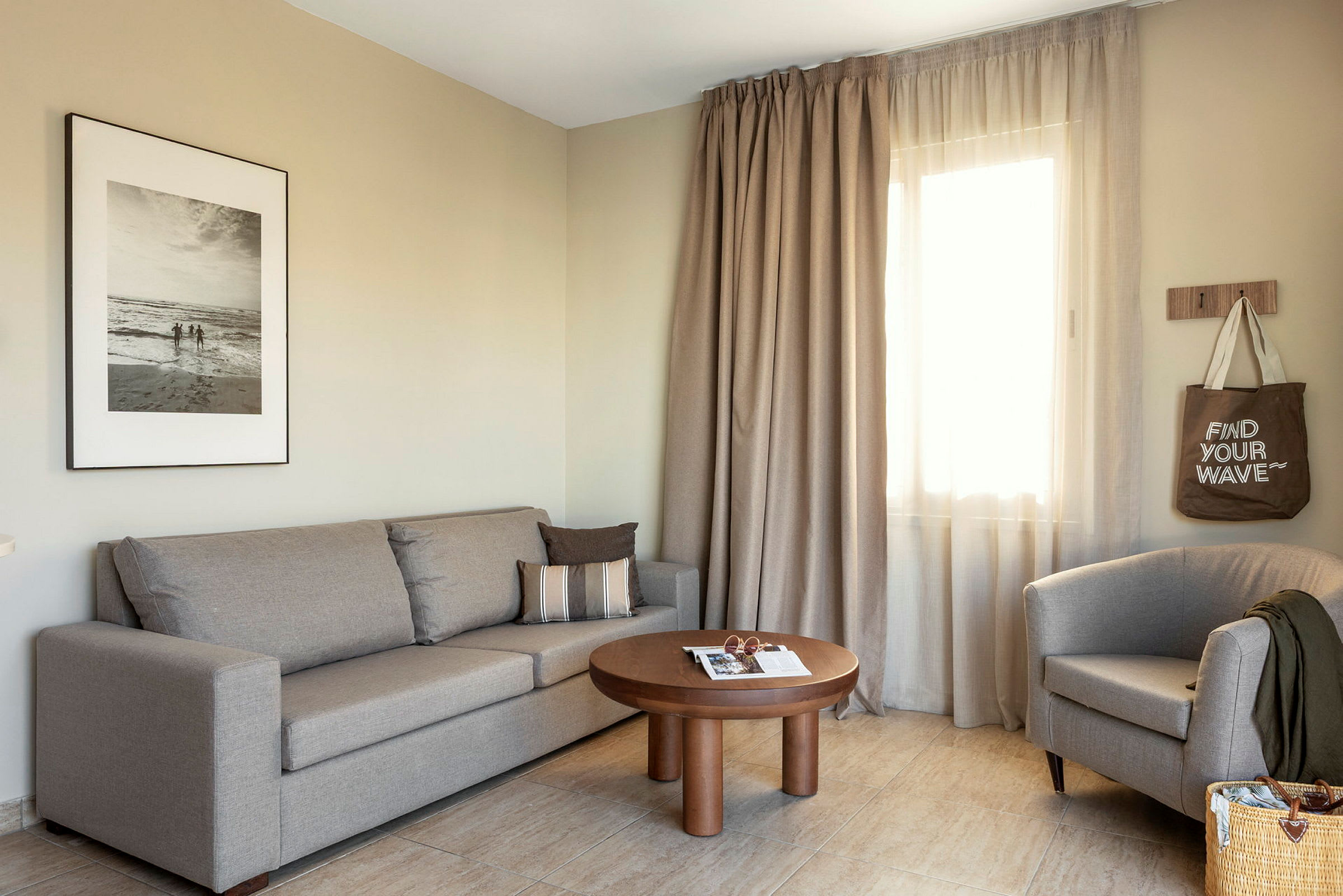 En stue med en beige sofa, en lænestol, et træbord og gardiner ved et vindue. (AI-genereret tekst)
