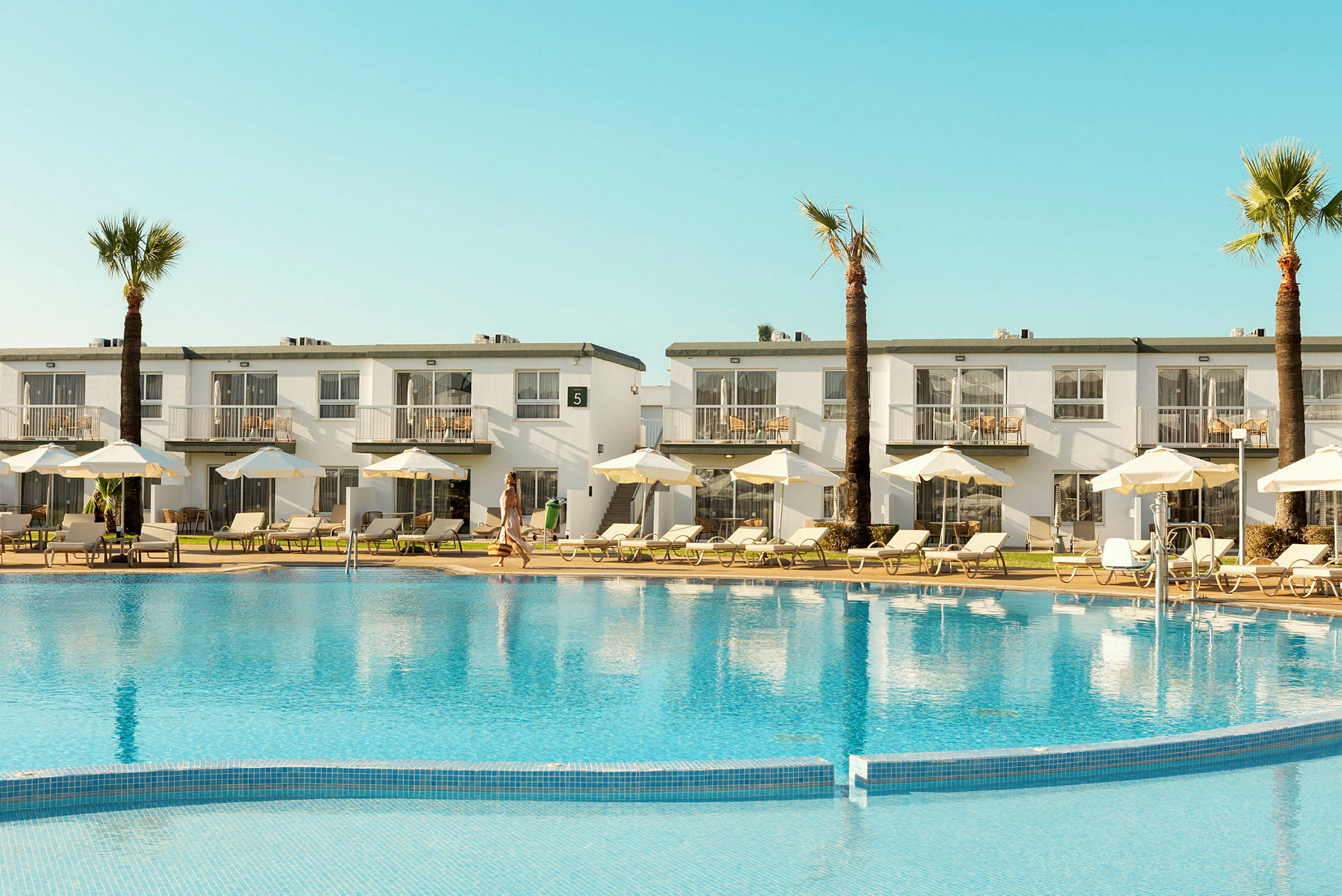 Hotelbygning med altaner og en pool foran, omgivet af solsenge og parasoller. (AI-genereret tekst)