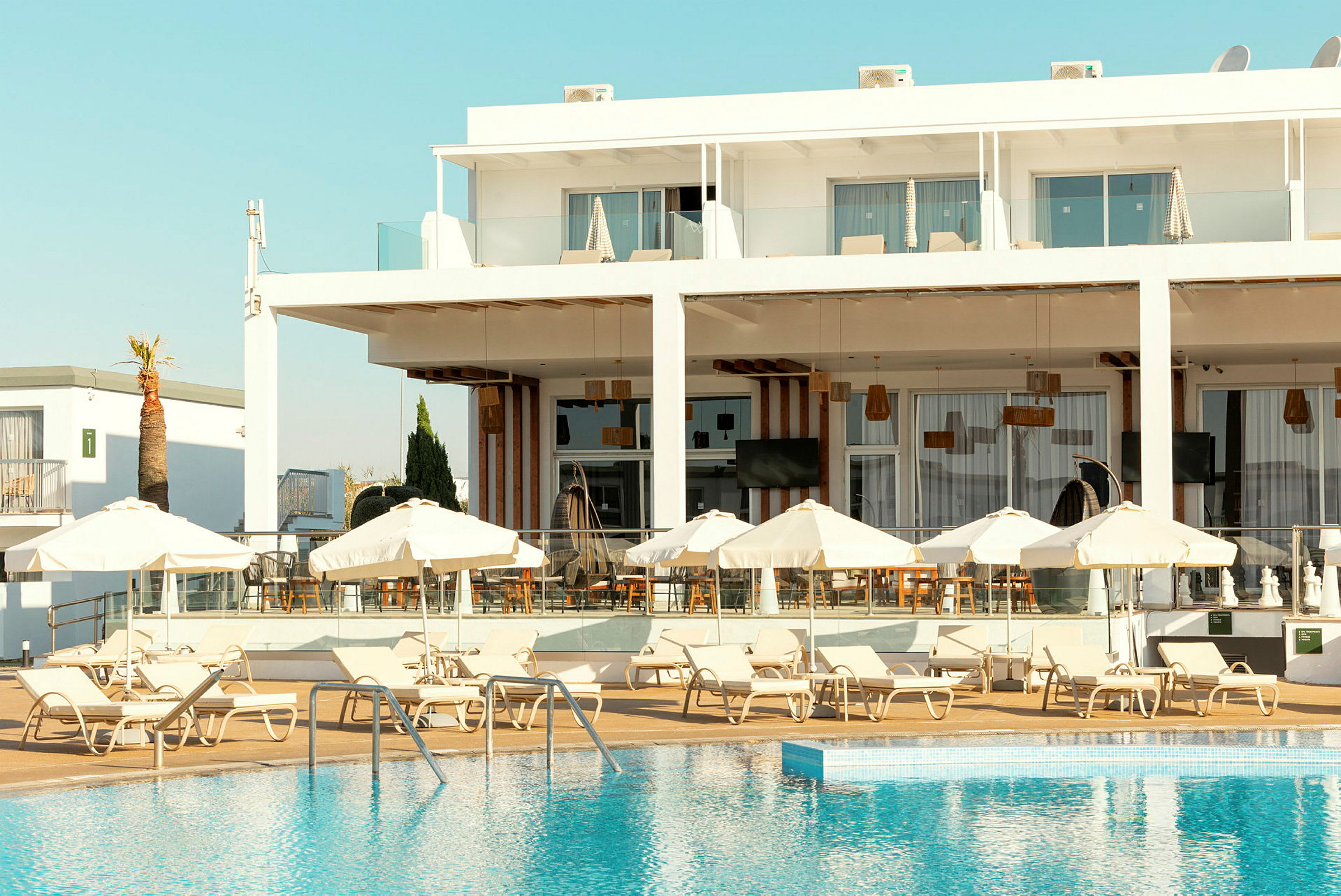 Billede af et hotel med liggestole og parasoller ved en pool. (AI-genereret tekst)