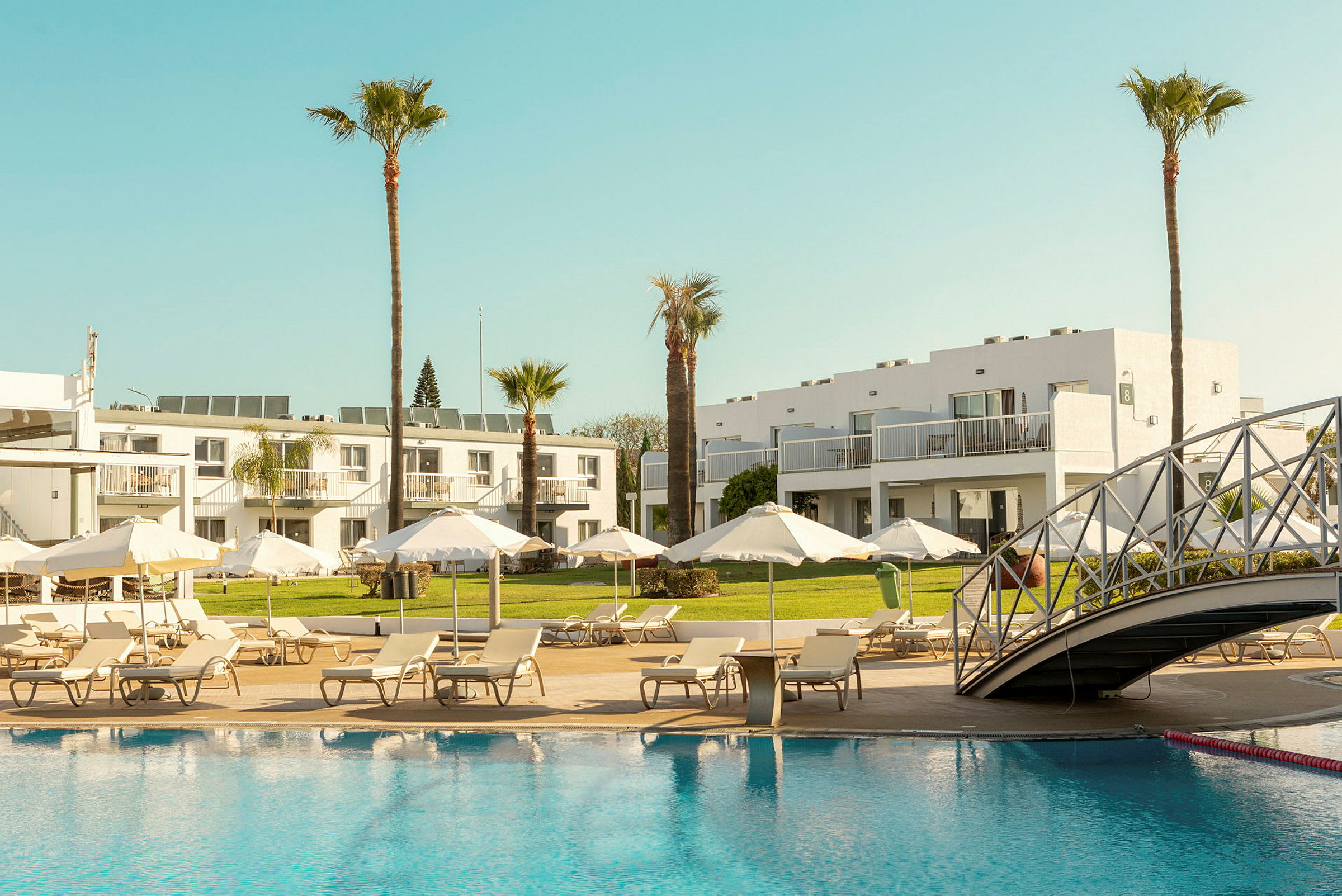 Hotelområde med pool, liggestole og parasoller samt hvide bygninger i baggrunden. (AI-genereret tekst)