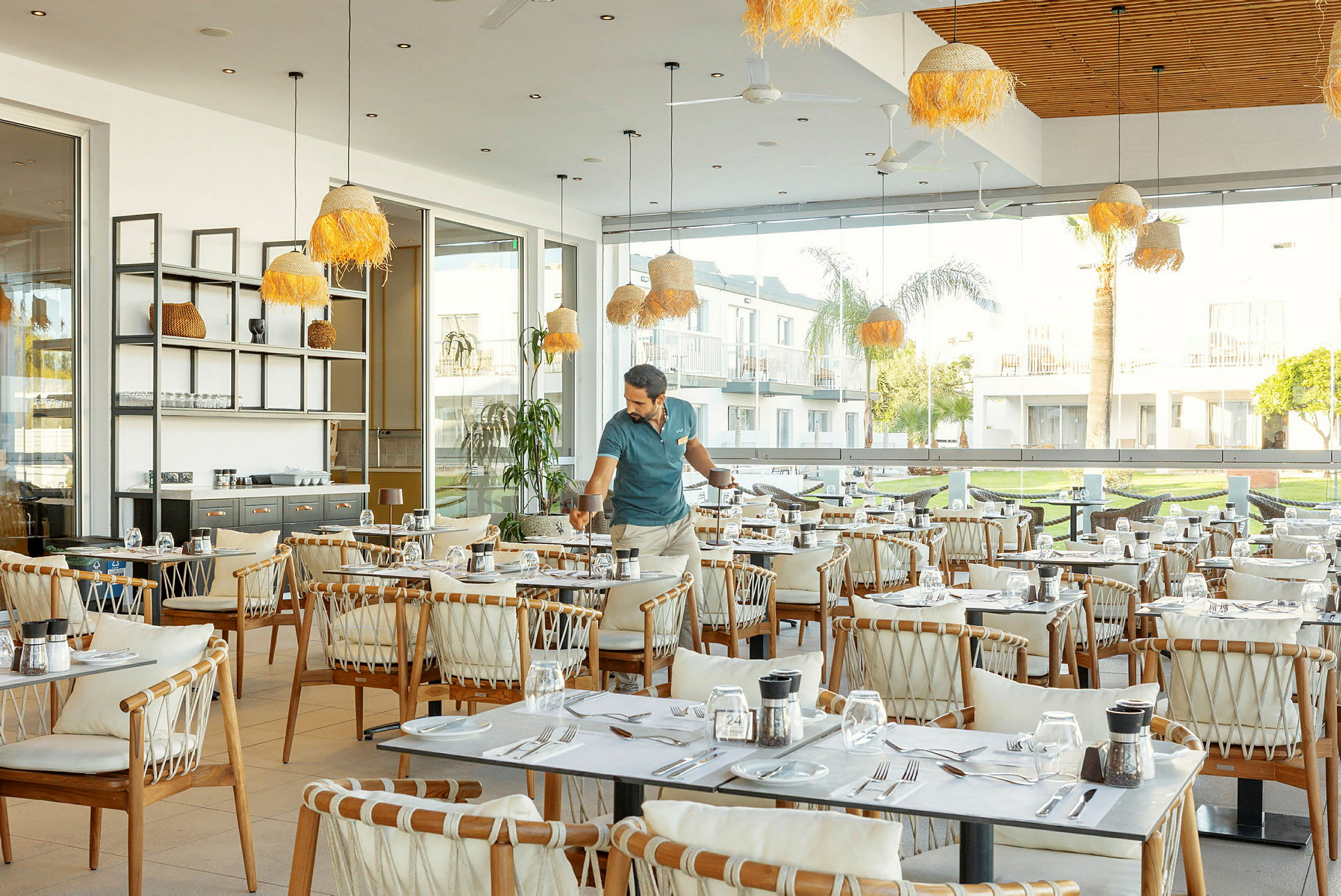 Restaurant med borde og stole samt store vinduer, der vender ud mod et grønt udendørsområde. (AI-genereret tekst)