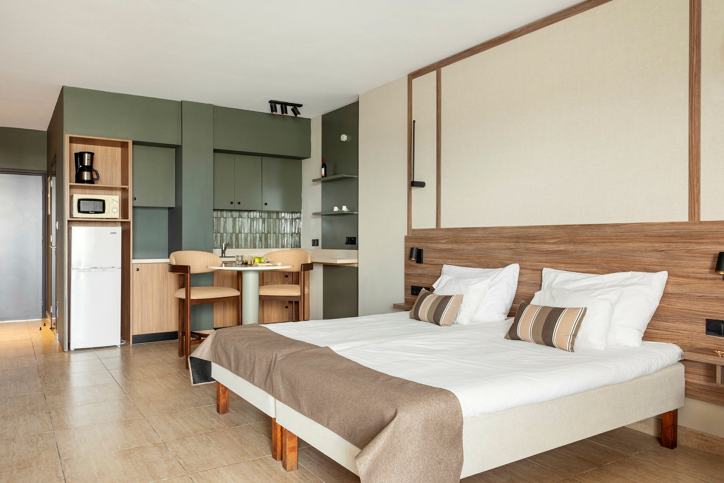 Indendørs billede af et lyst og moderne hotelværelse med en seng, bord, stole og køkkenområde. (AI-genereret tekst)