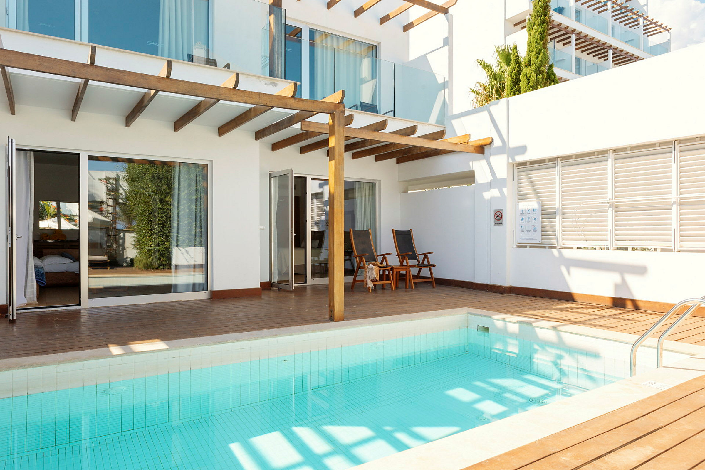 En mindre swimmingpool med en træterrasse og moderne hvide bygninger i baggrunden. (AI-genereret tekst)