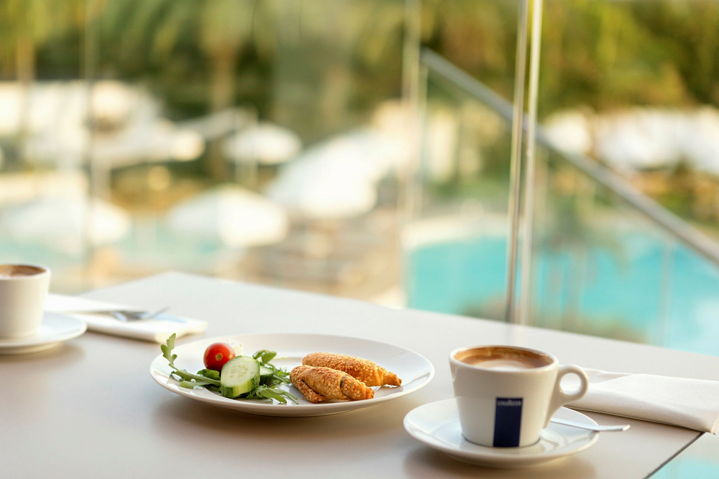 Morgenmadsscene med kaffe og en tallerken med grøntsager og bagværk, baggrund med udsigt over en pool. (AI-genereret tekst)