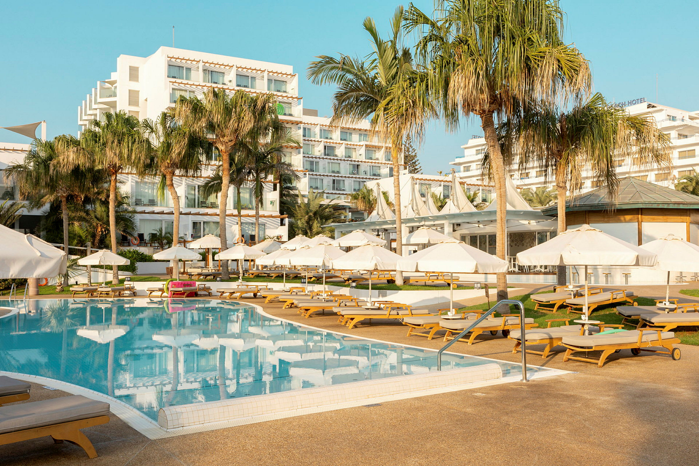 Billede af en hotelpool med liggestole og parasoller, omgivet af palmer og hotelbygninger. (AI-genereret tekst)