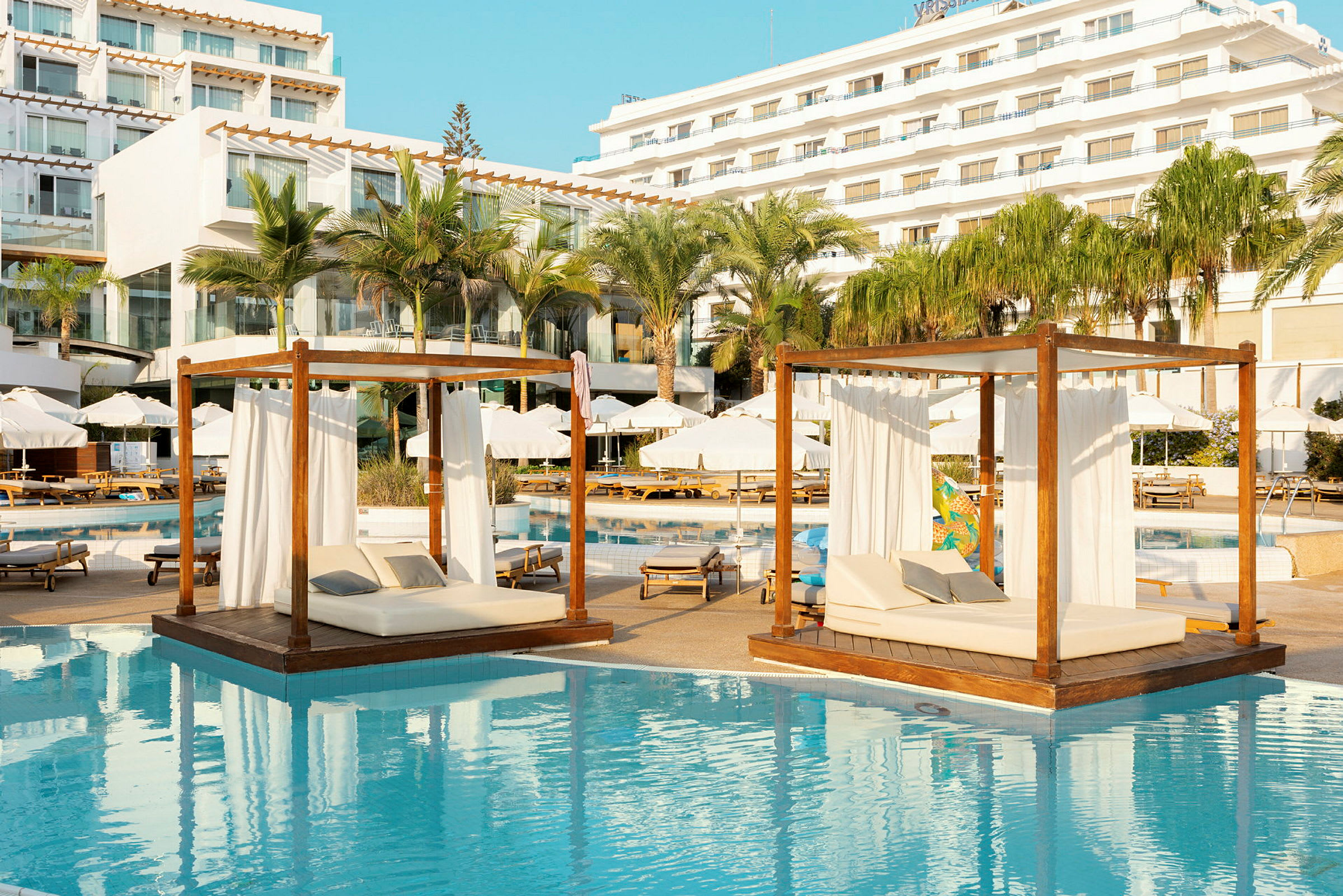 Liggende dagsenge med hvide gardiner over vand ved en solrig hotelpool. (AI-genereret tekst)