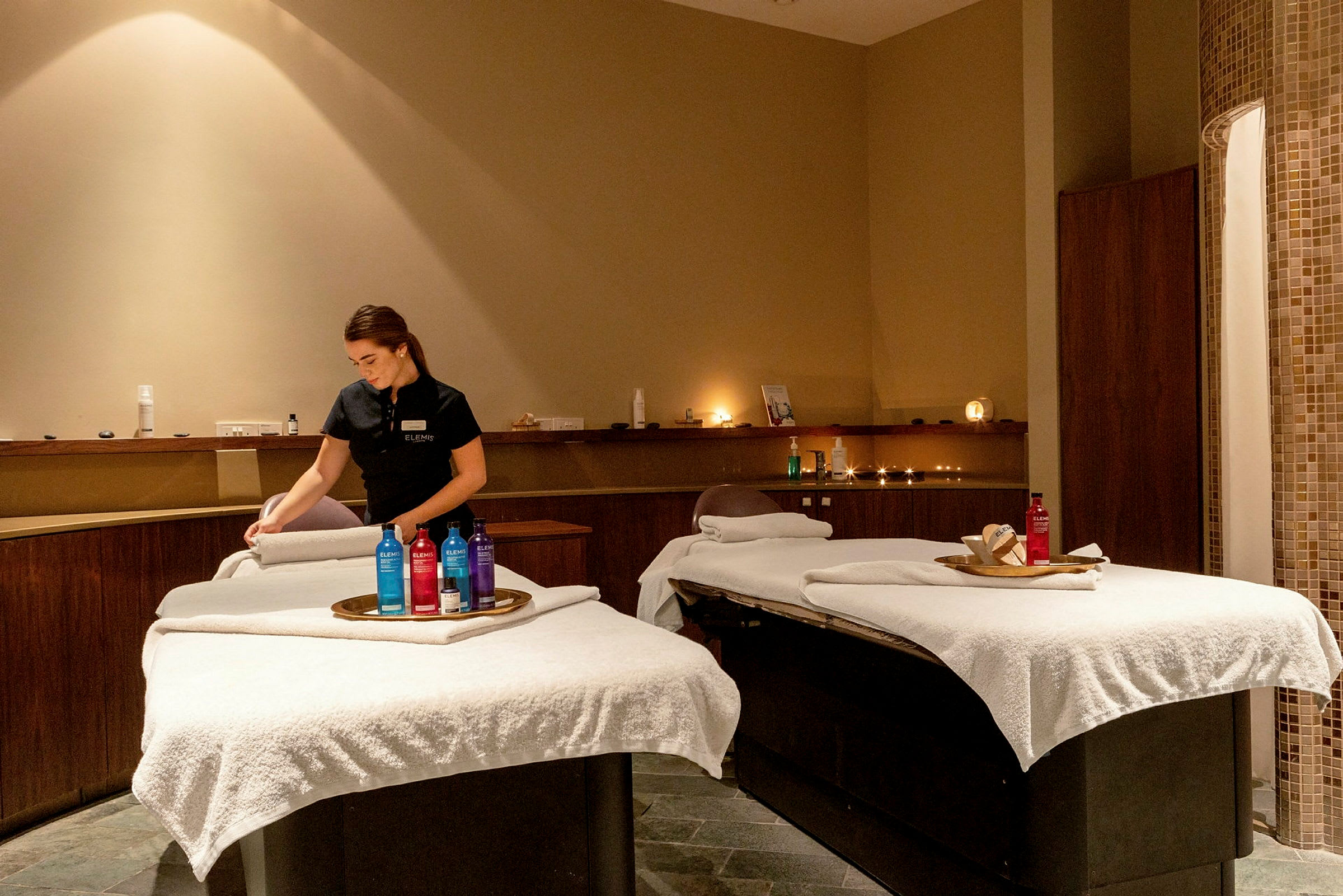 Et spa-rum med to massagestole dækket med hvide duge og produkter på bakker. (AI-genereret tekst)