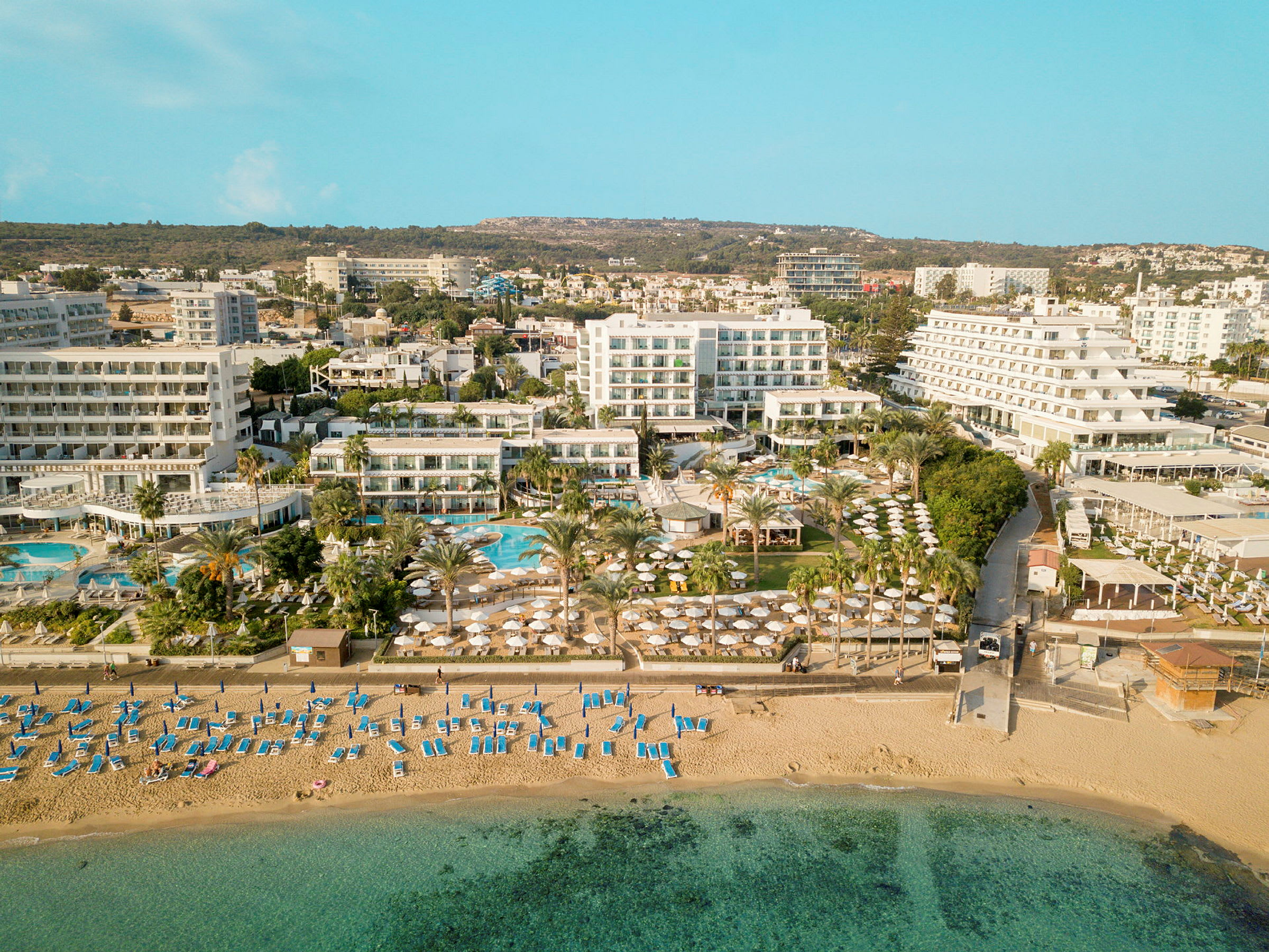 Luftfoto, der viser en strand med liggestole og parasoller samt hotelbygninger og poolområde i baggrunden. (AI-genereret tekst)