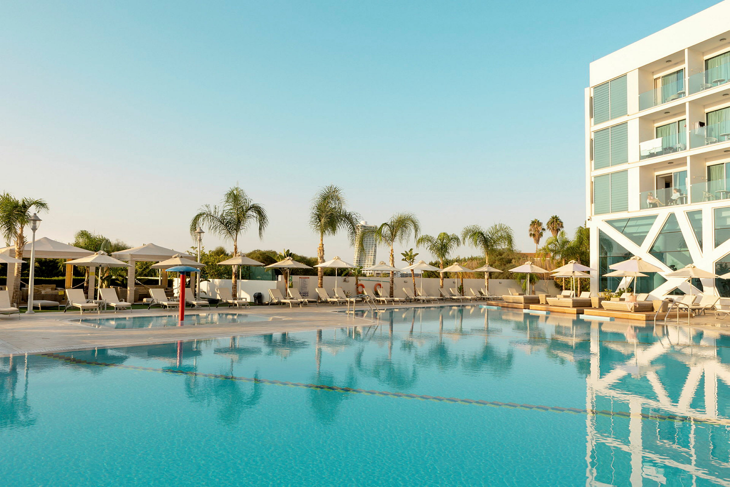 En udendørs swimmingpool omgivet af liggestole og parasoller samt et hotel i baggrunden. (AI-genereret tekst)