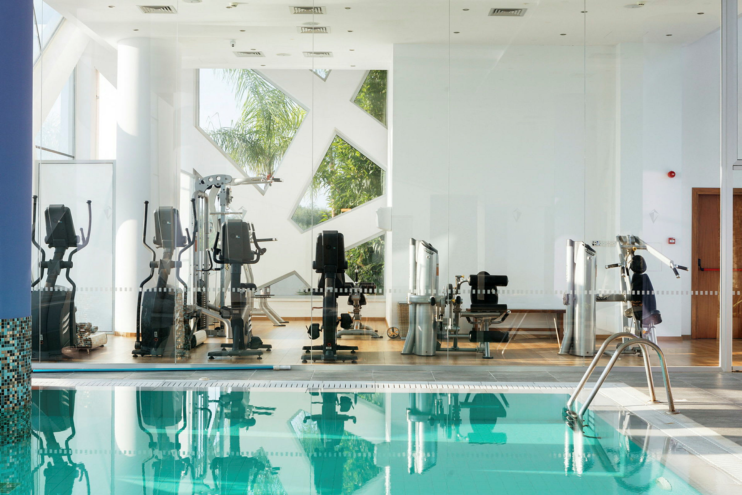 Billede af fitnessudstyr bag glasvægge nær en pool. (AI-genereret tekst)
